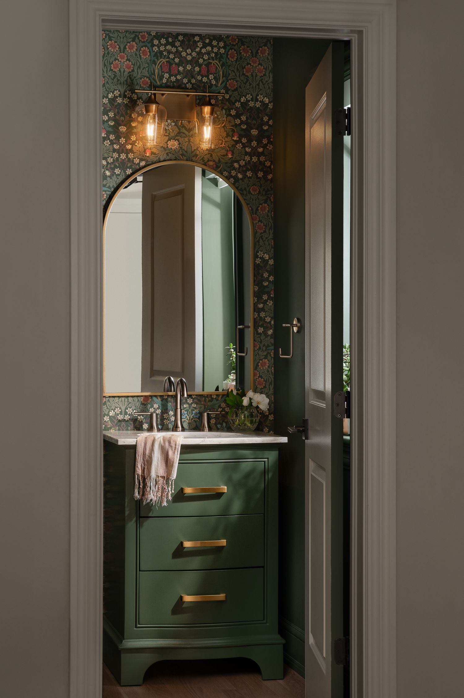 Elegant powder room featuring dark floral wallpaper, arched mirror, and green vanity with brass hardware.