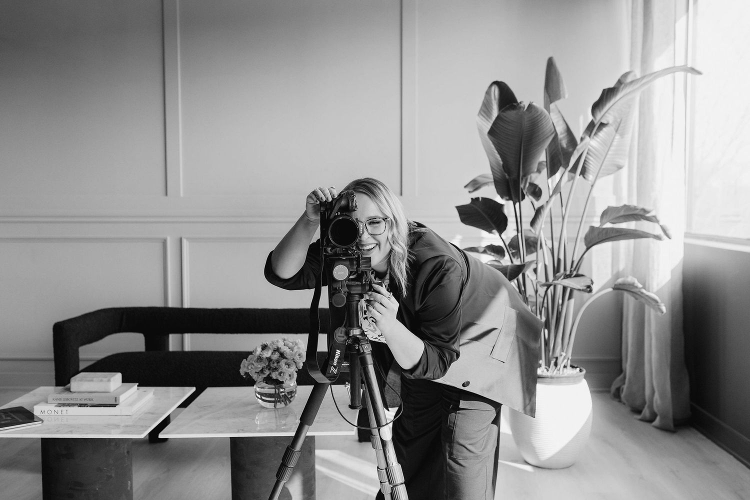 Black and white photo of a photographer using a tripod-mounted camera in an interior setting with a potted plant nearby.