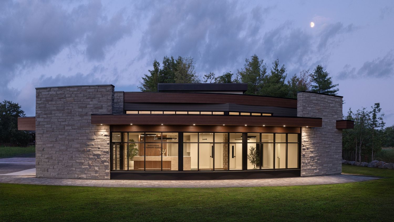 Modern stone and glass building exterior illuminated at dusk with lush green trees and a crescent moon in the background.