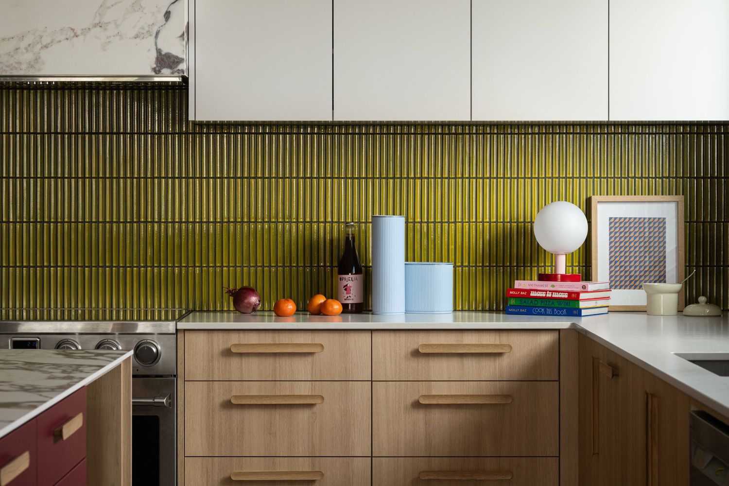 Modern kitchen with olive green vertical tile backsplash, wood cabinets, white countertops, and decorative items including books and fruit.