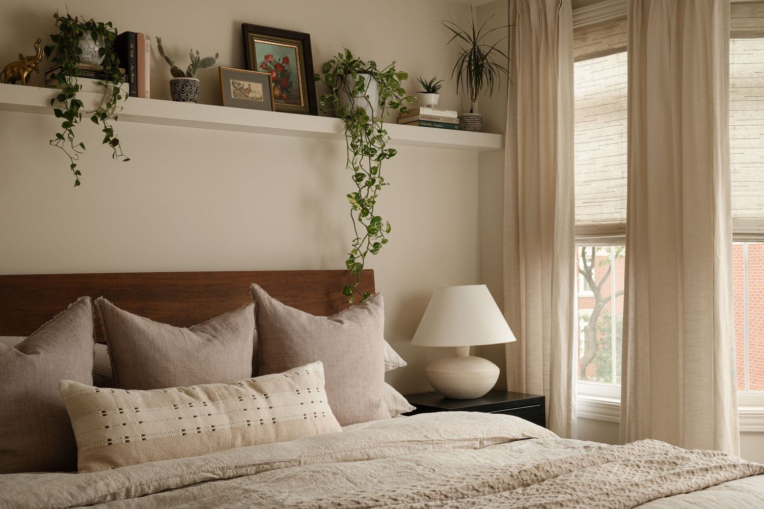 Cozy bedroom with wood headboard, neutral pillows, white lamp, sheer curtains, and lush green plants on a floating shelf.
