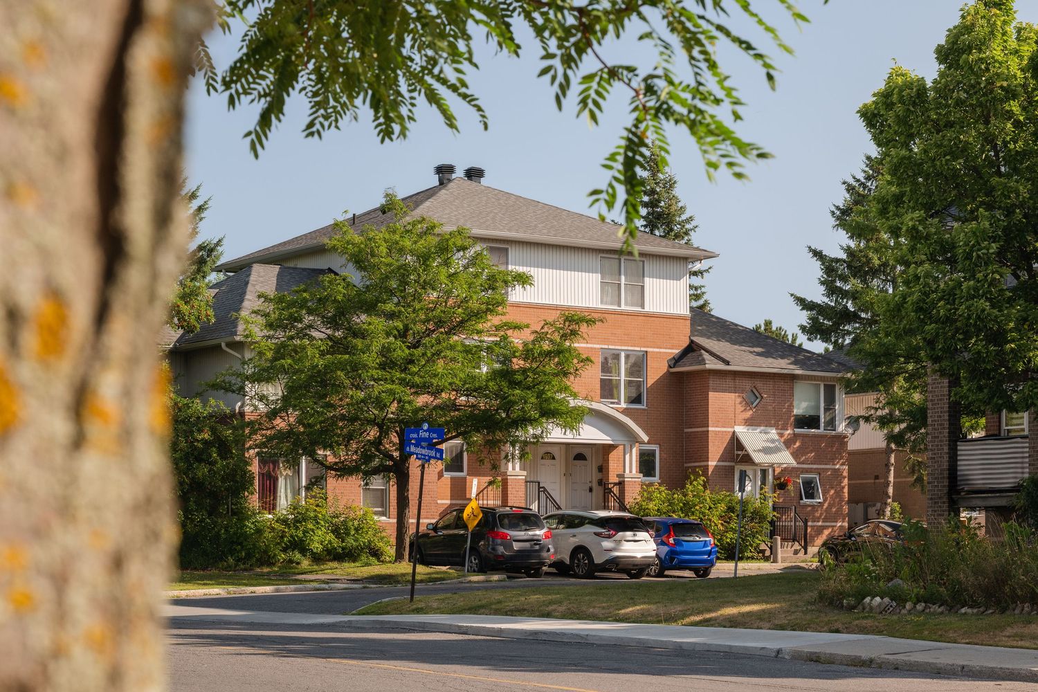 Peaceful suburban street view with brick houses, parked cars and mature trees lining the residential neighborhood.