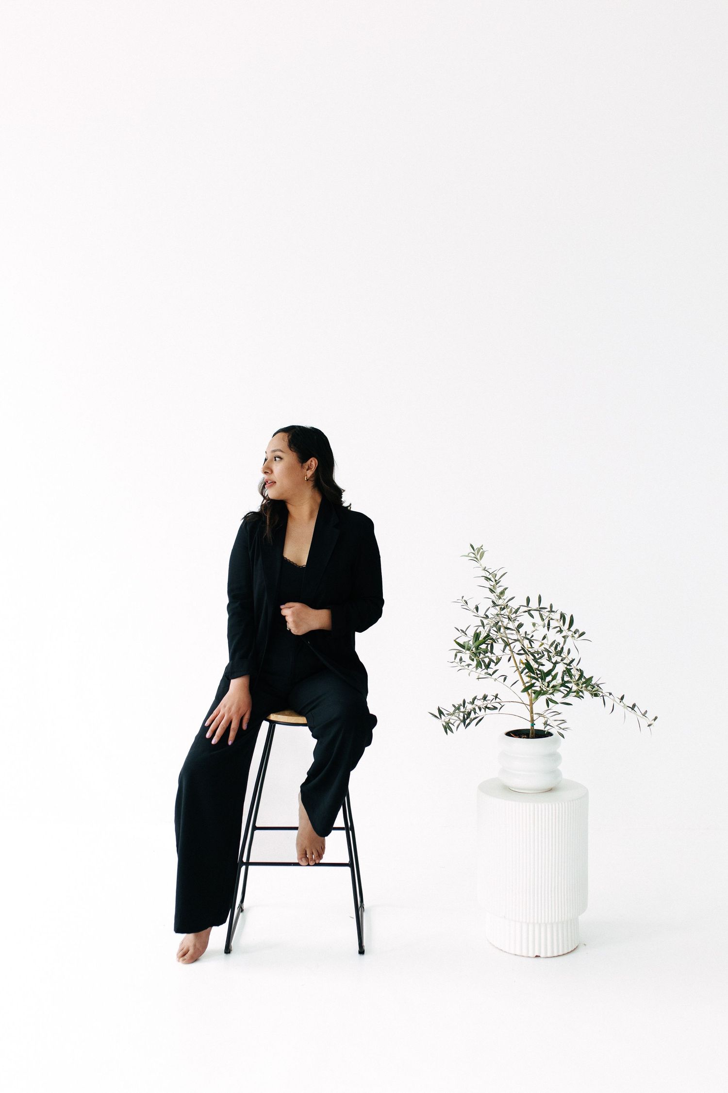 Elegant studio portrait featuring model in dark clothing on stool beside white ceramic planter.