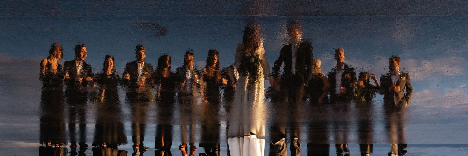The colorful reflection of a wedding party standing in a line as shown by the puddle on a New England hangar runway .