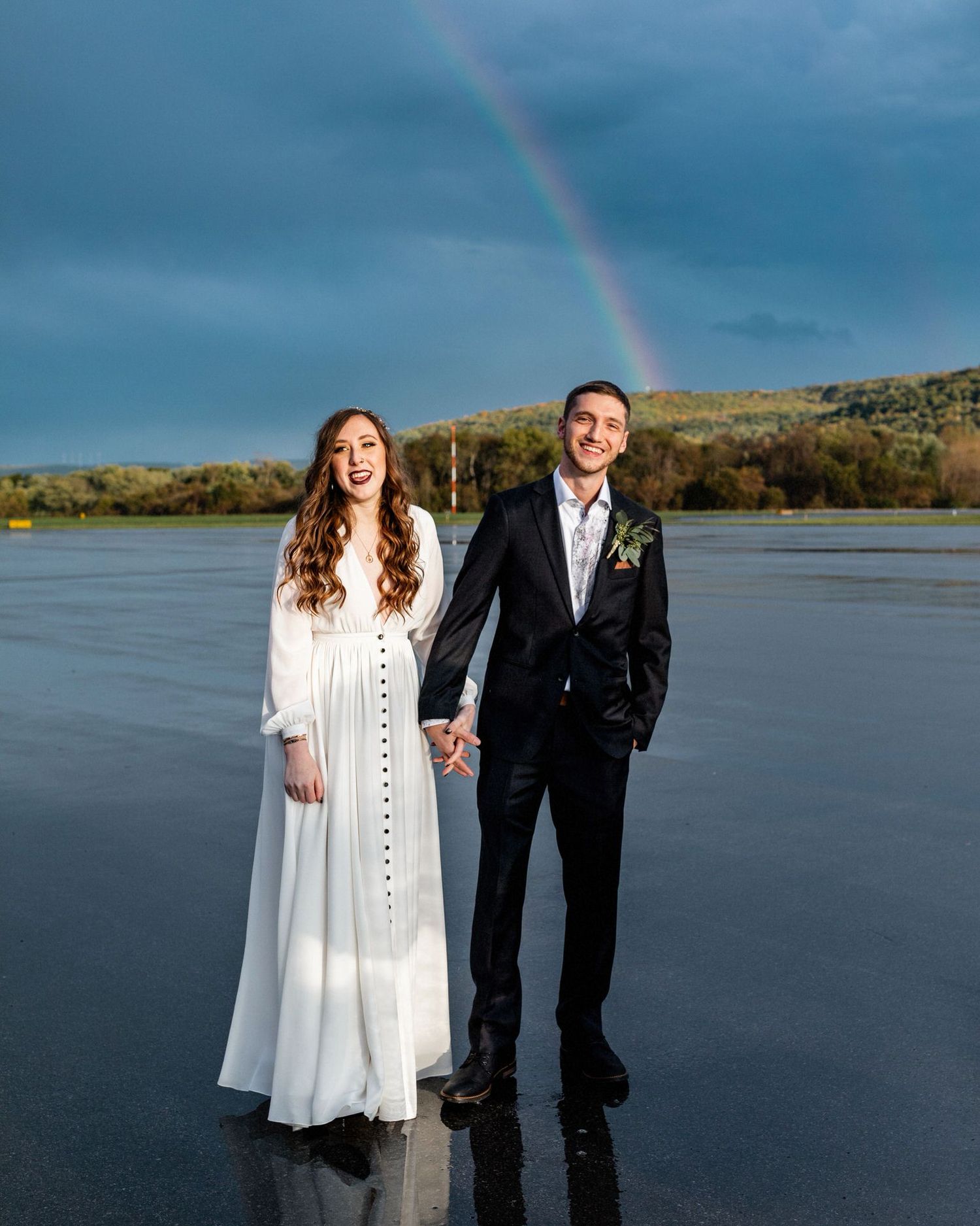 A New England eloping couple holds hands and stands on a small airport runway in their wedding attire in front of a blue sky and rainbow.