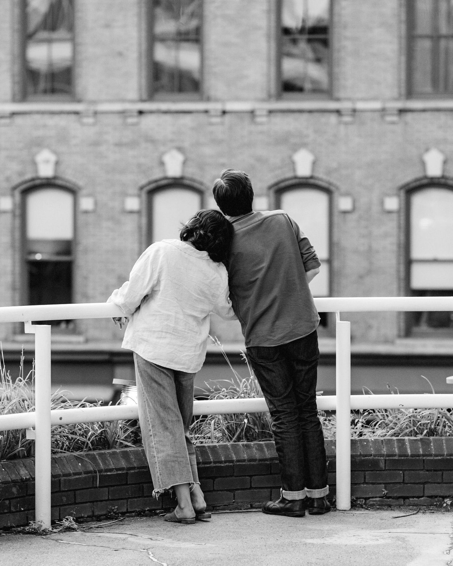 A black and white photo of a couple taken from behind as they stand against a railing and cuddle in Troy, NY.