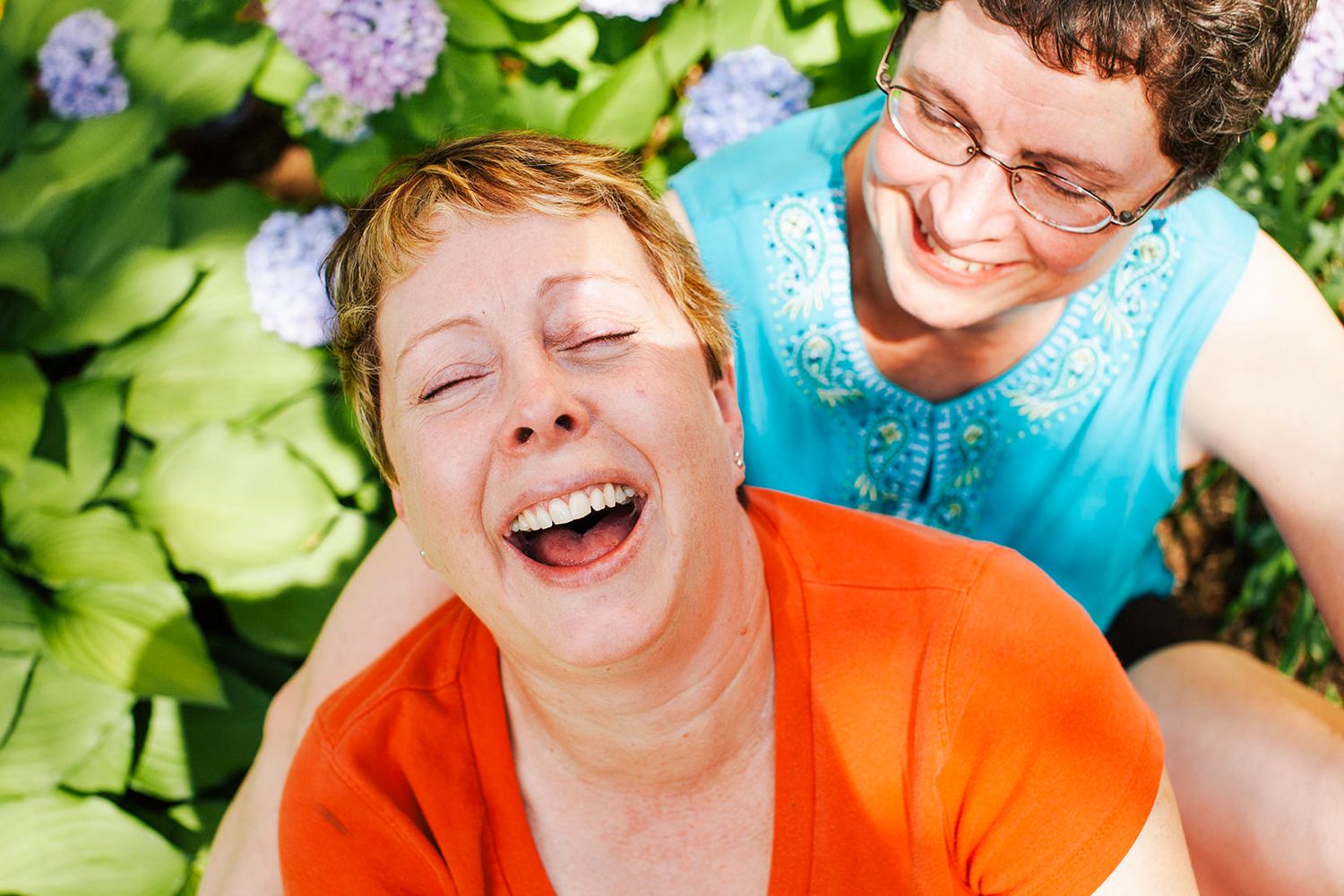 A candid photo of a couple of smiling white women holding each other in a New England garden while throwing their heads back in laughter.