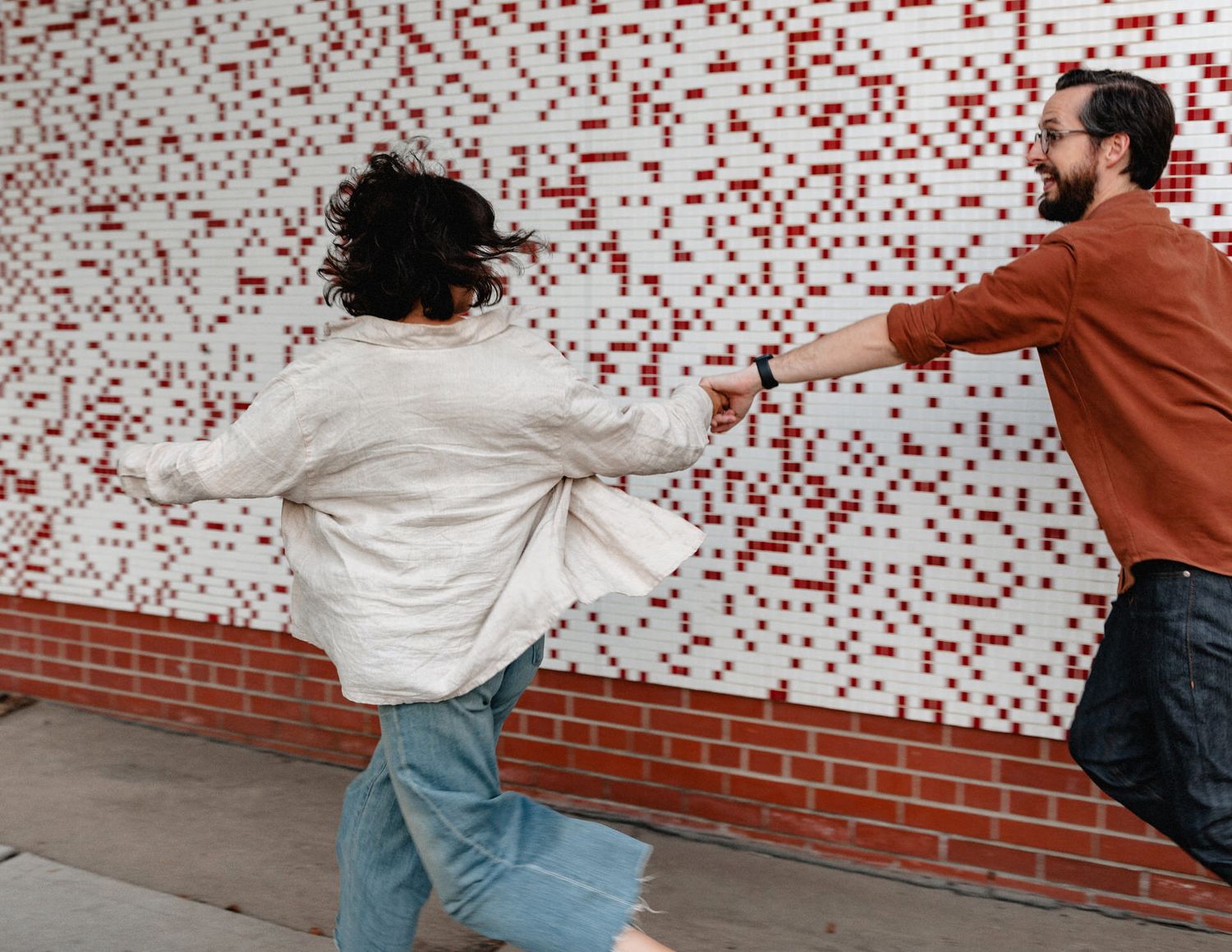 A happy couple laughs and holds hands as they run in front of a red and white tiled building in Troy, NY.