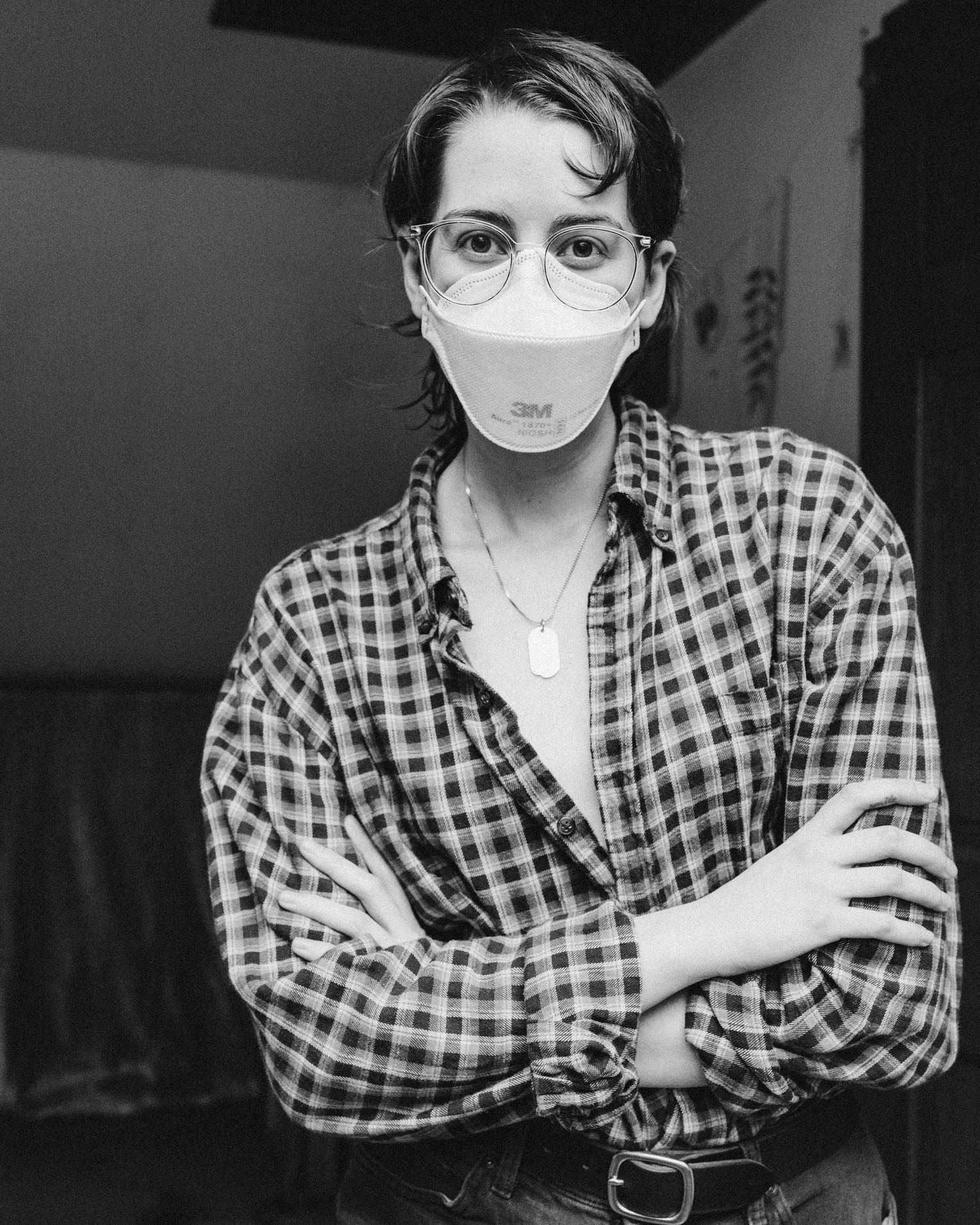 A black and white photo of Ro Tatchi wearing an N95 respirator and glasses with their arms crossed in front of their plaid shirt.