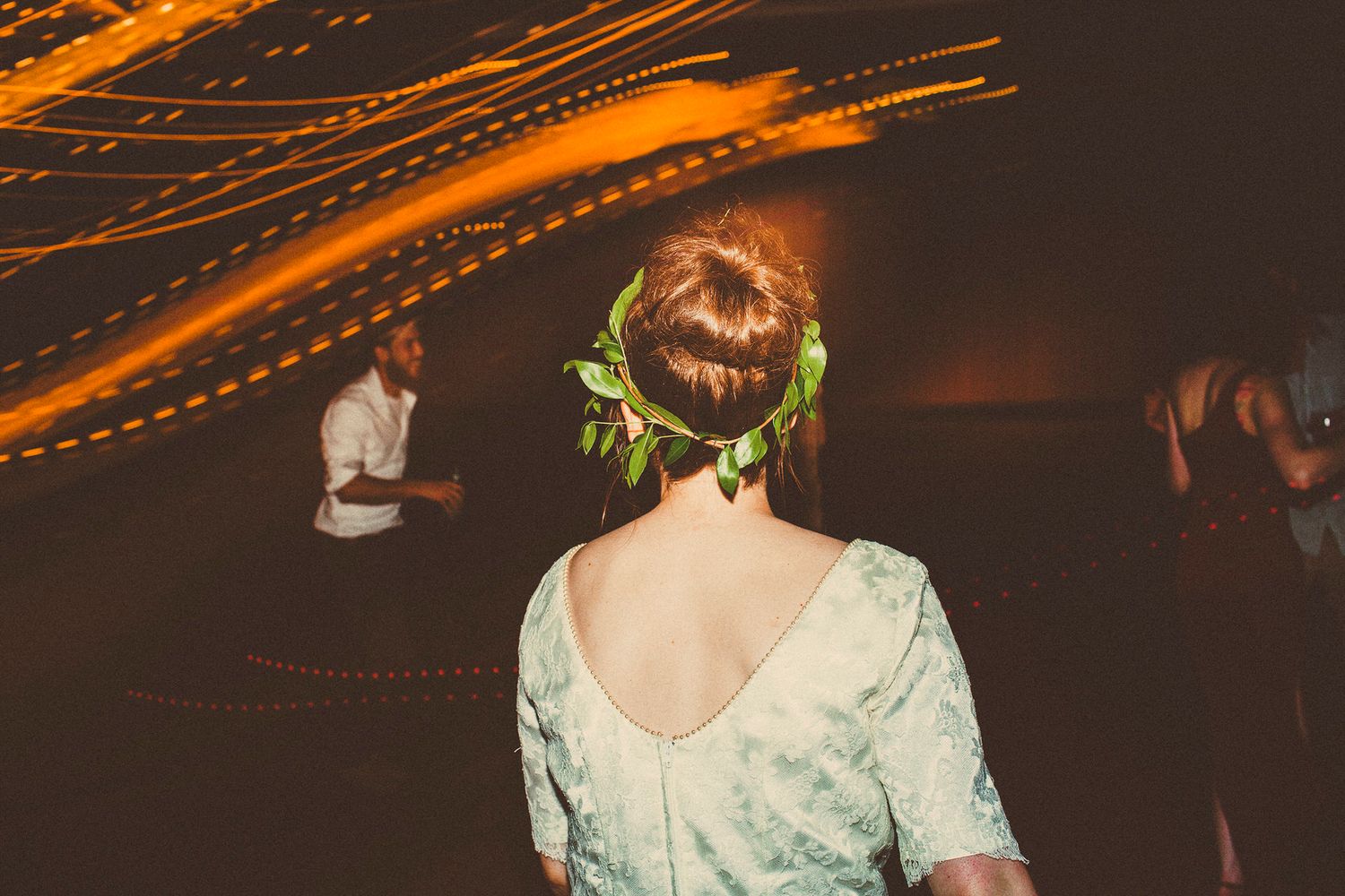 A film photo of a bride wearing a string of leaves as a veil and walking onto the dance floor of golden orange lights streaming across.