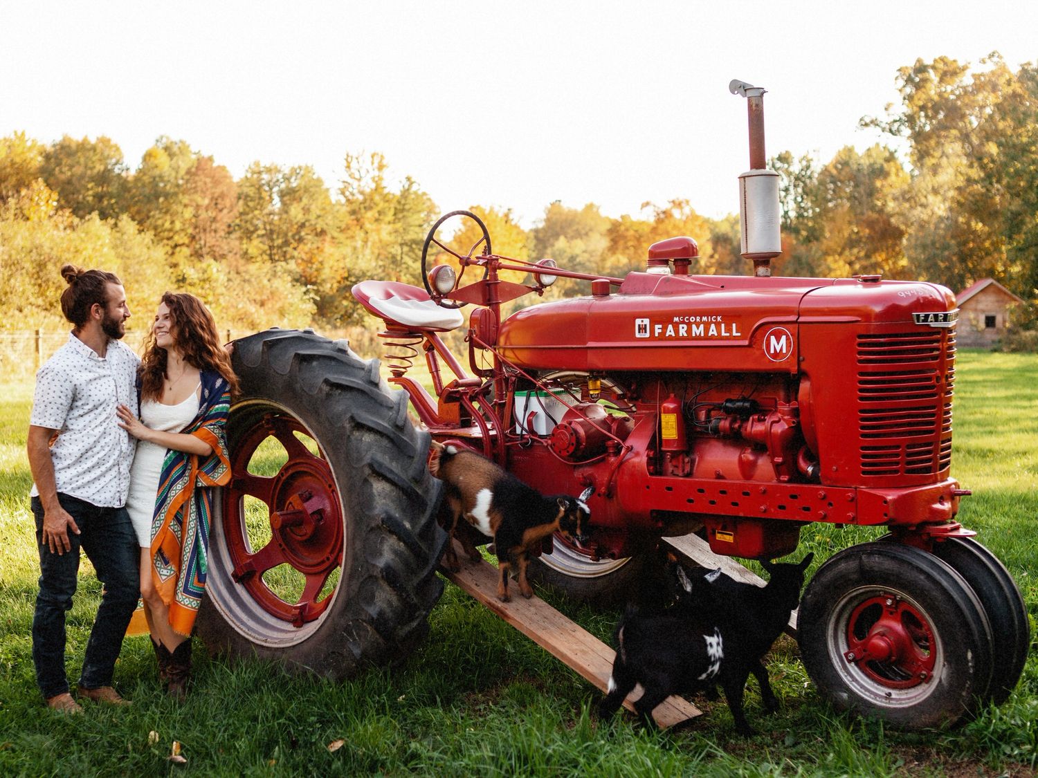 A white couple hugging in front of a red tractor at sunset on a New England farm.
