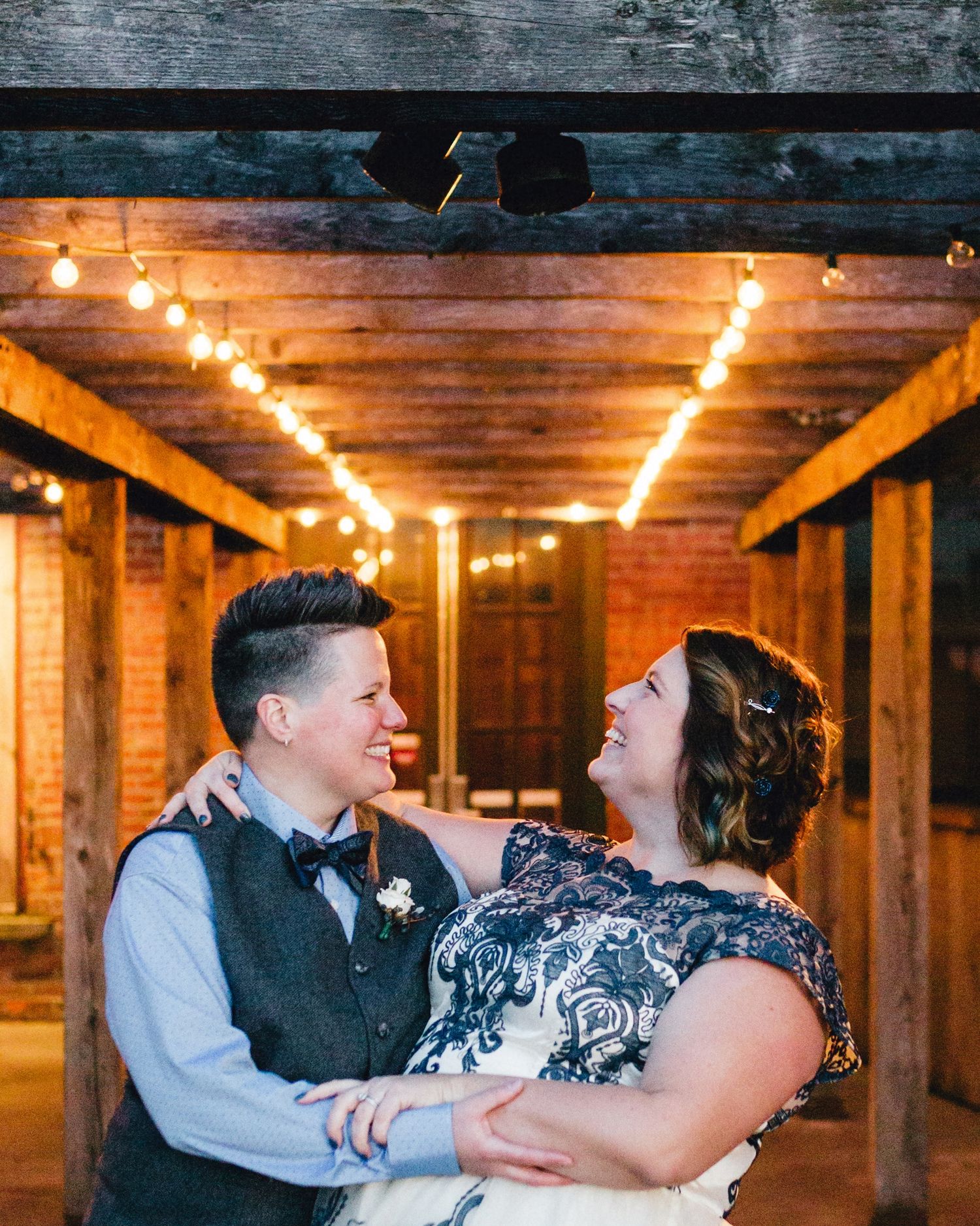 A just married lesbian couple holds each other and smiles as they dip under the warm hue of outdoor lights. They wear masc and femme outfits