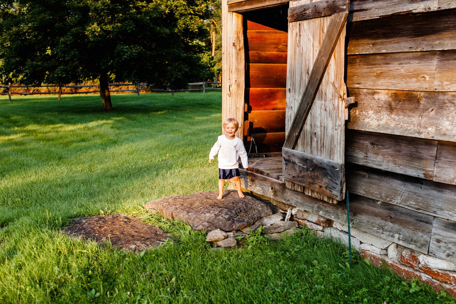 A young blonde boy stands on a stone in golden light at a New England barn entrance and smiles surrounded by green grass.