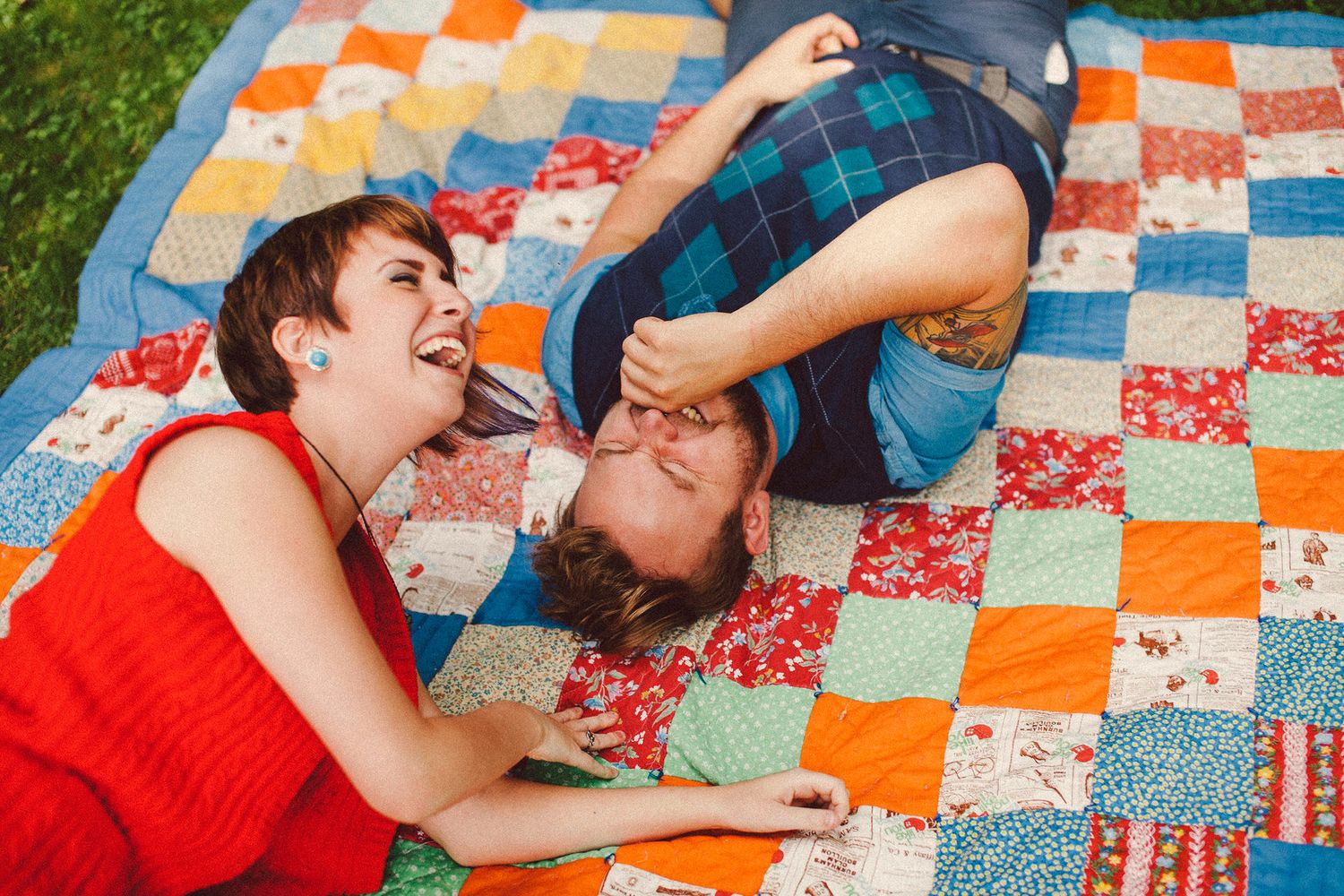 A white couple wearing vibrant outfits laughs with big smiles while rolling on a bright colorful picnic quilt in a New England park.