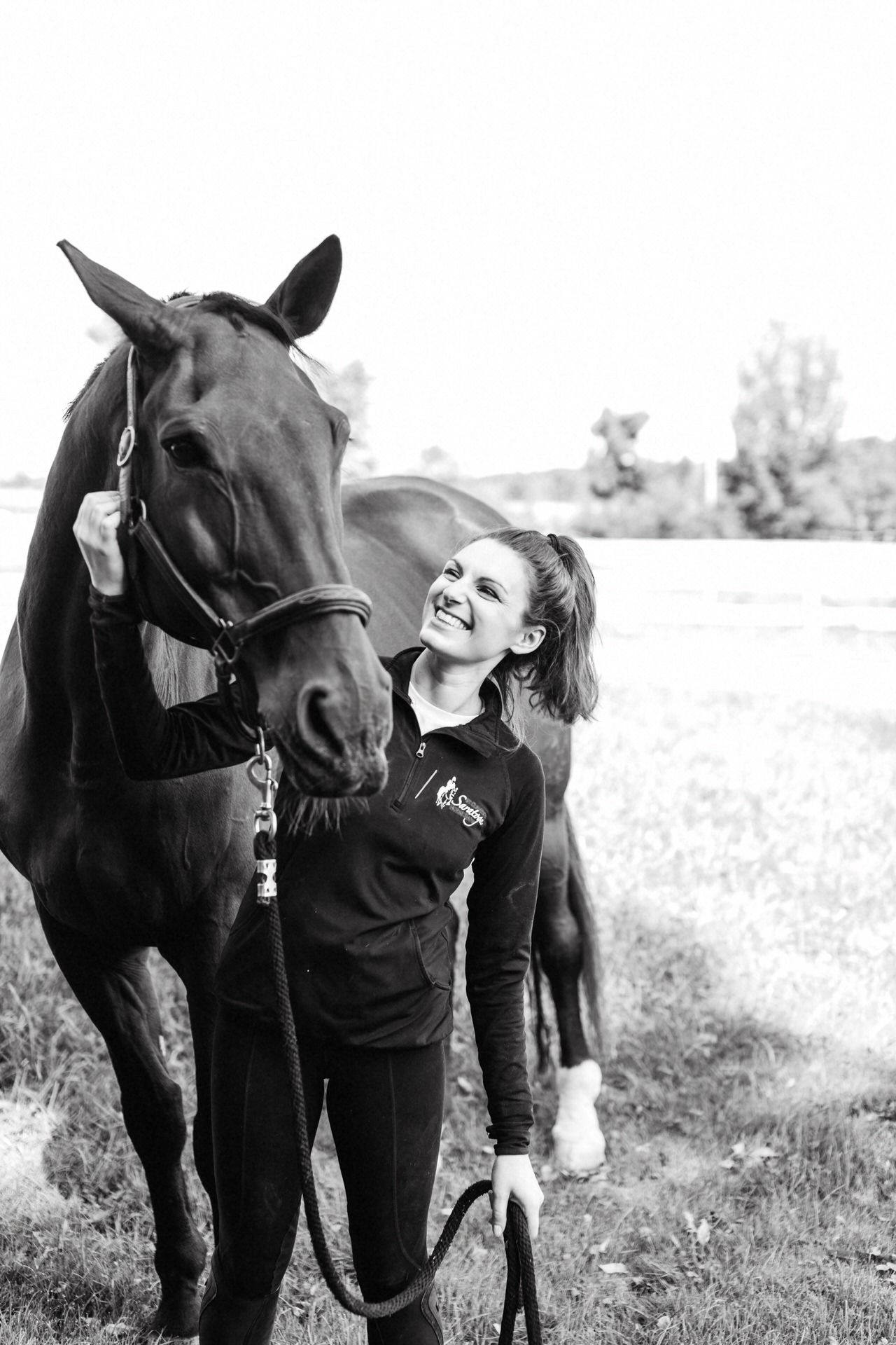 A black and white image of a smiling white woman and her horse as they walk around a New England field.