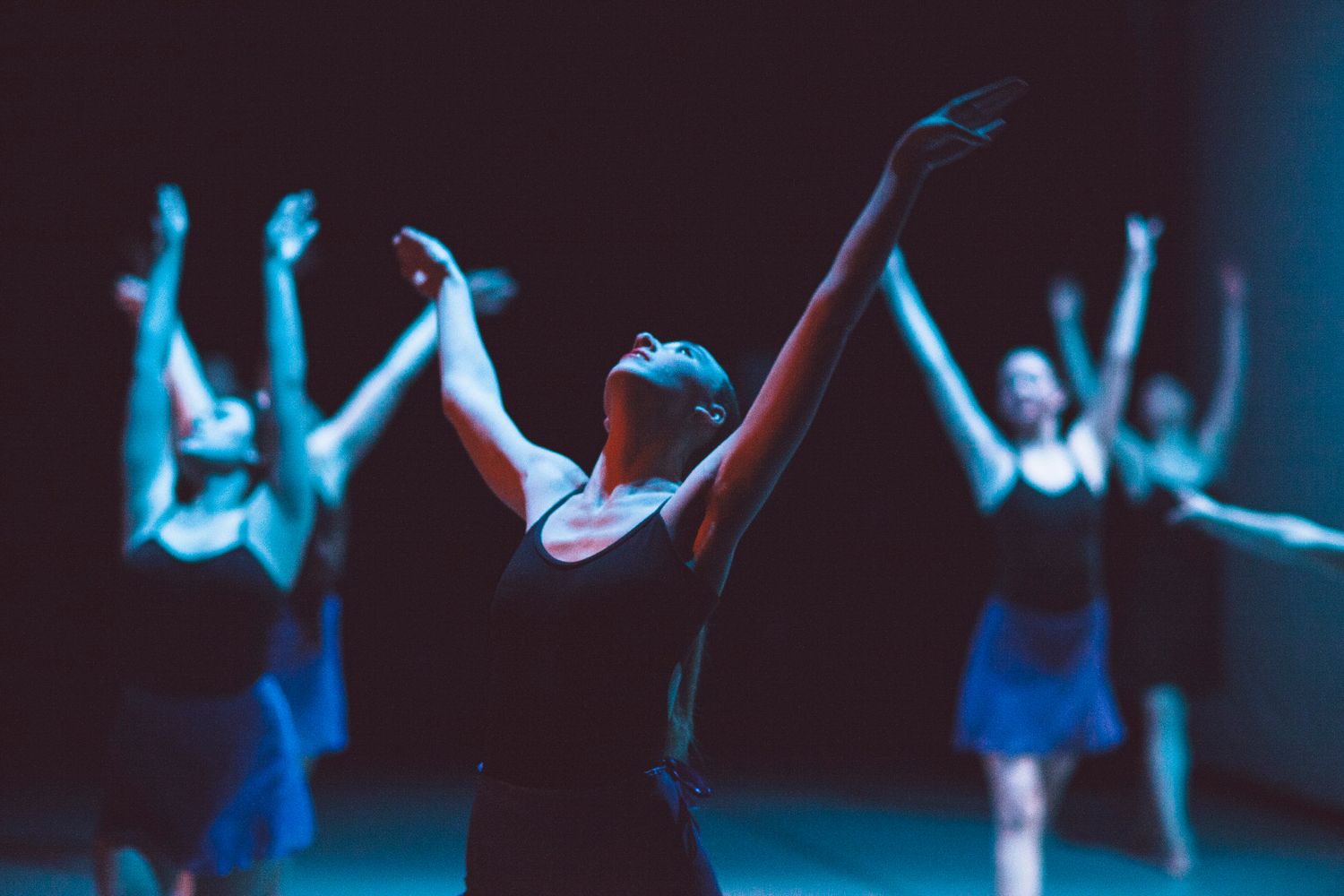 5 dance performers in black leotards reaching up above their heads on a stage under teal lights.