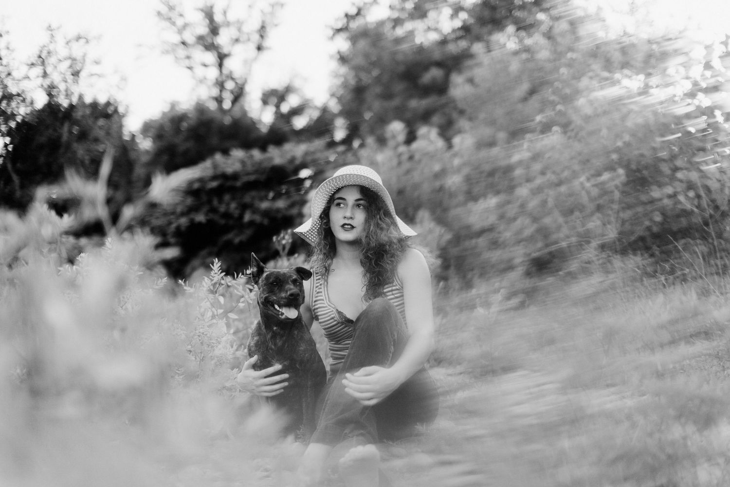 A black and white portrait of a woman in a big sun hat and her brindle pit bull in the brush on the side of the river in New England.