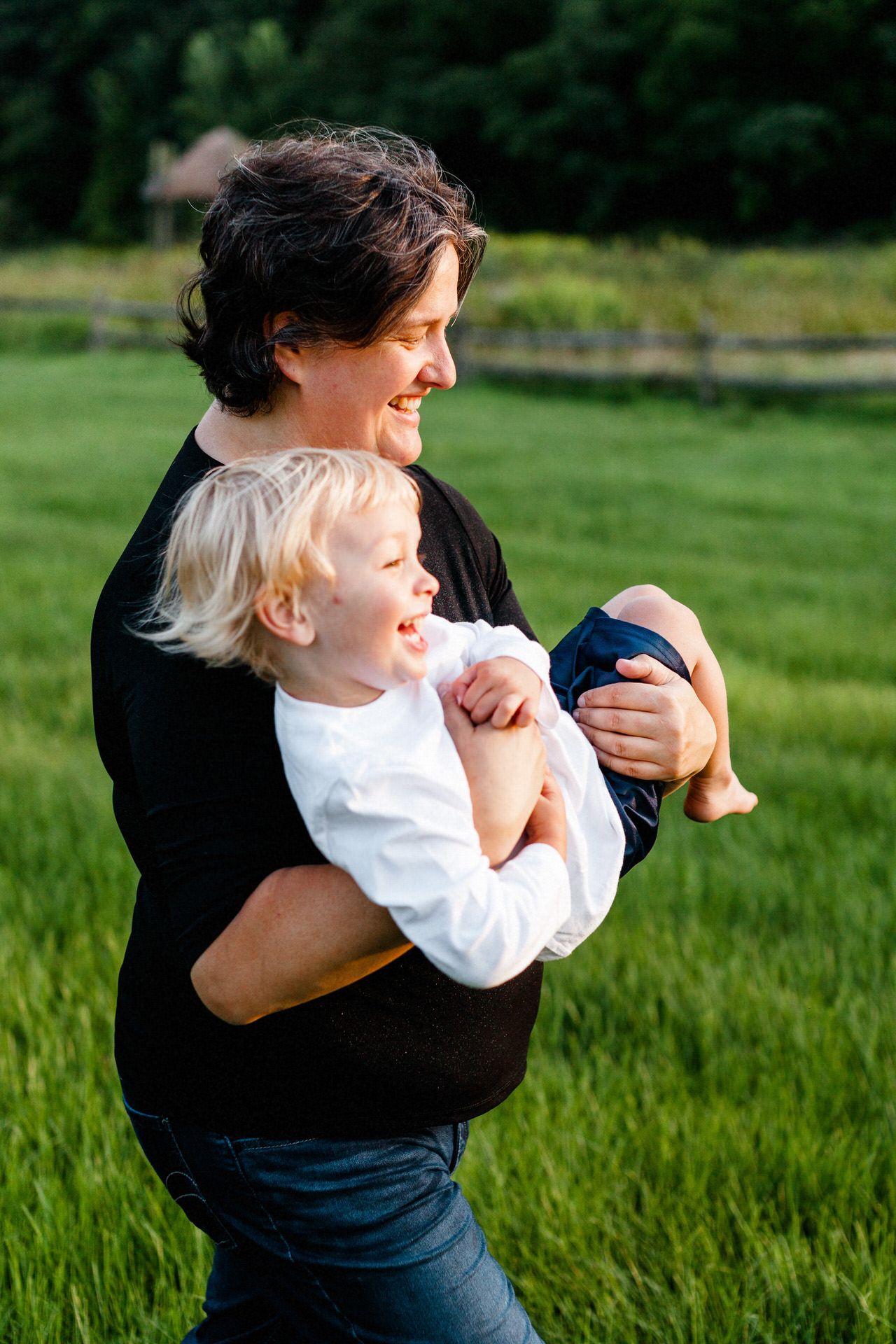 A brown-haired white mother holds her blonde-haired son in her arms as they giggle and run across a Vermont field of green grass.