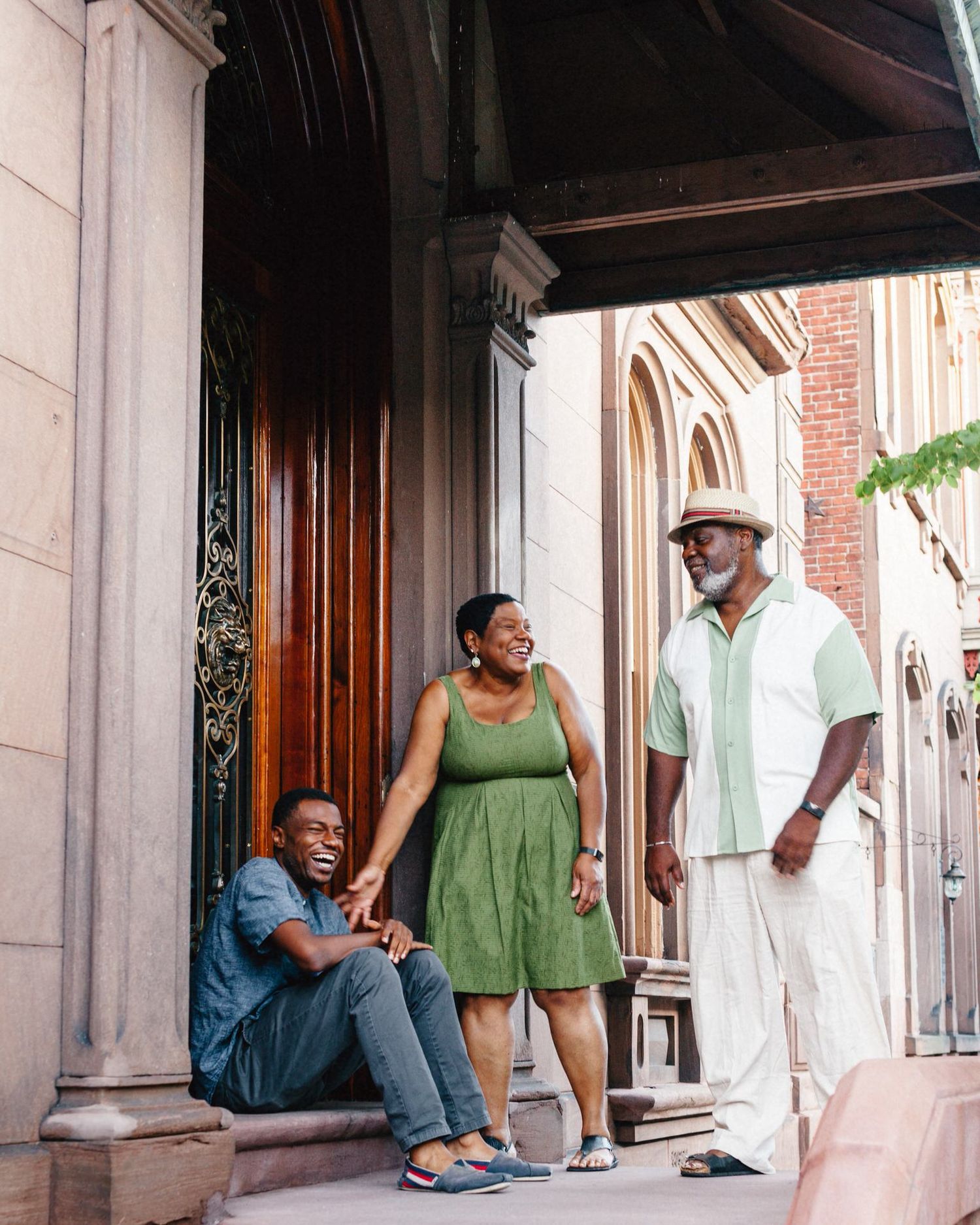 A classy Black family of three laughs together on a brownstone porch in Troy, New York. The father and mother stand and their adult son sits