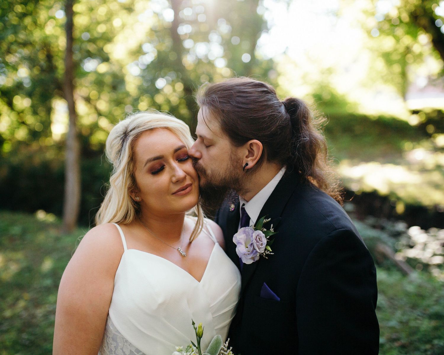 A New England elopement couple stands in the forest backlit. The long-haired groom kisses the blonde bride on the cheek as she smiles.