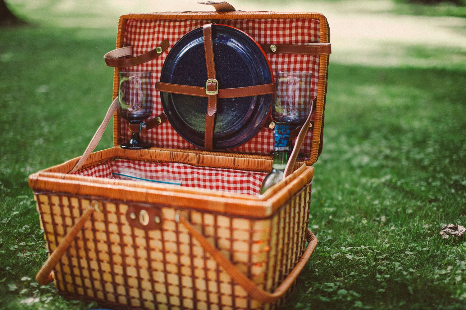 A wicker picnic basket with red gingham fabric lining the interior. It sits on the grass in Vermont holding blue enamel dishes for a picnic.