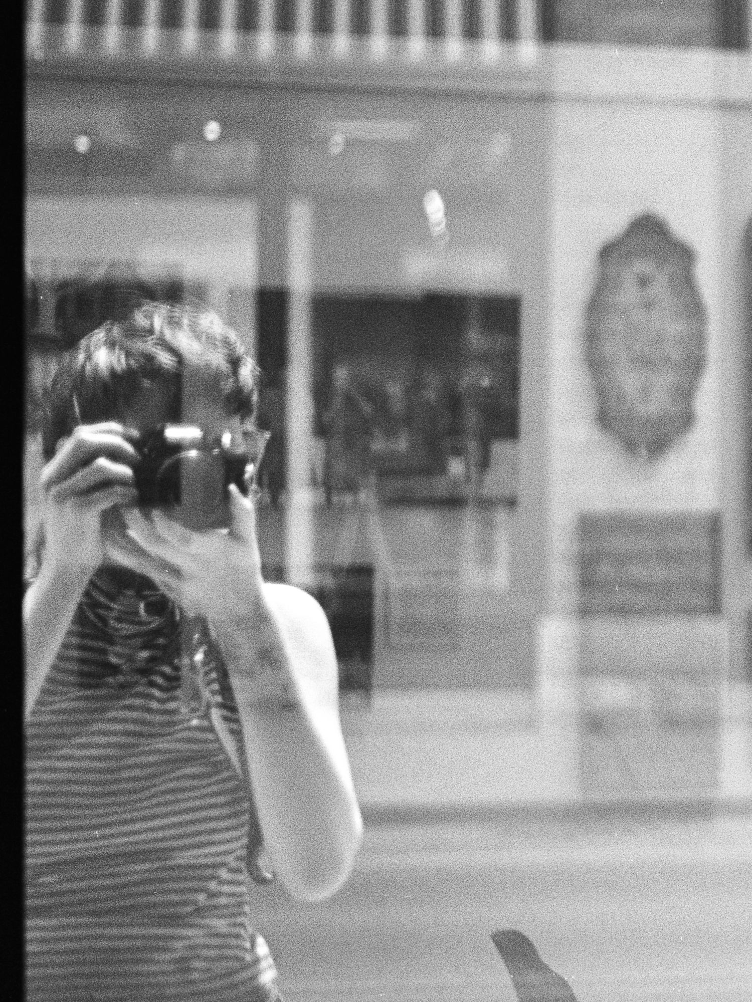A black and white film photo of Ro Tatchi taking a photograph into the window of a Middlebury Vermont art shop.
