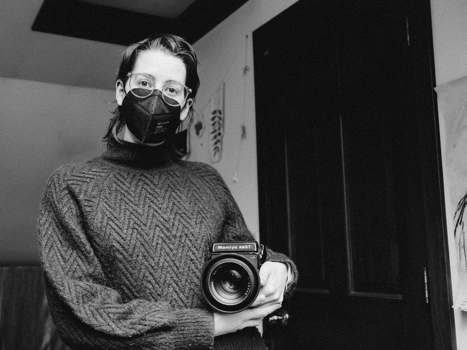 A black and white photograph of Ro Tatchi wearing a black KN95 and holding a medium format film photo. They are smiling behind the mask.