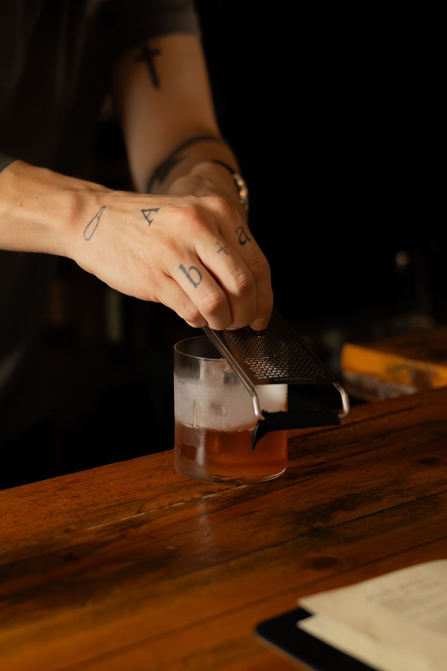 Hands preparing a cocktail on a wooden bar surface with strainer and mixing glass.