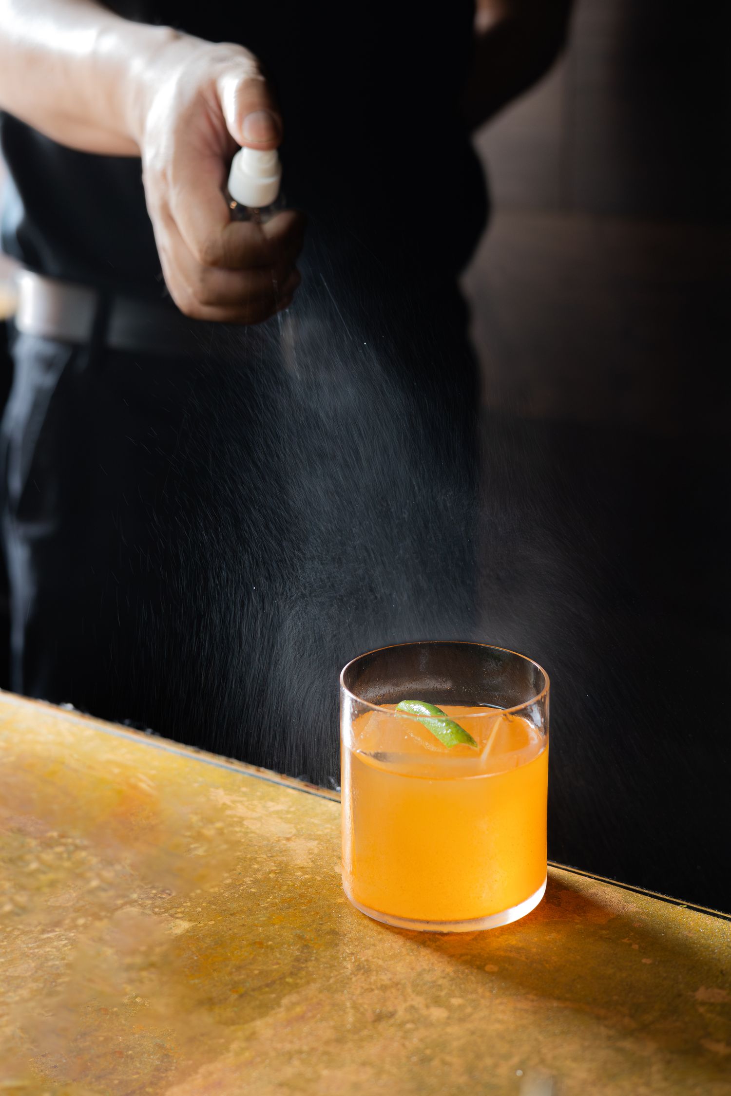 Bartender spraying mist over orange cocktail on golden bar top.