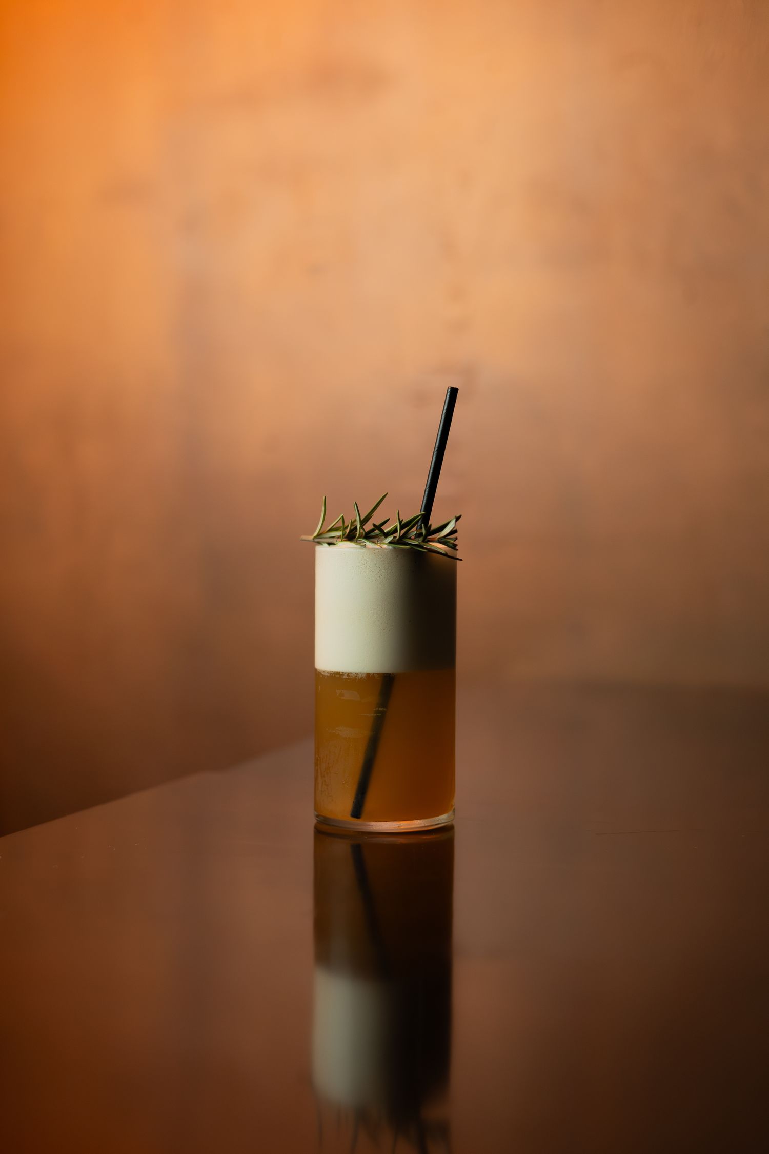 A layered cocktail with foam top garnished with rosemary served in a tall glass against an orange background.