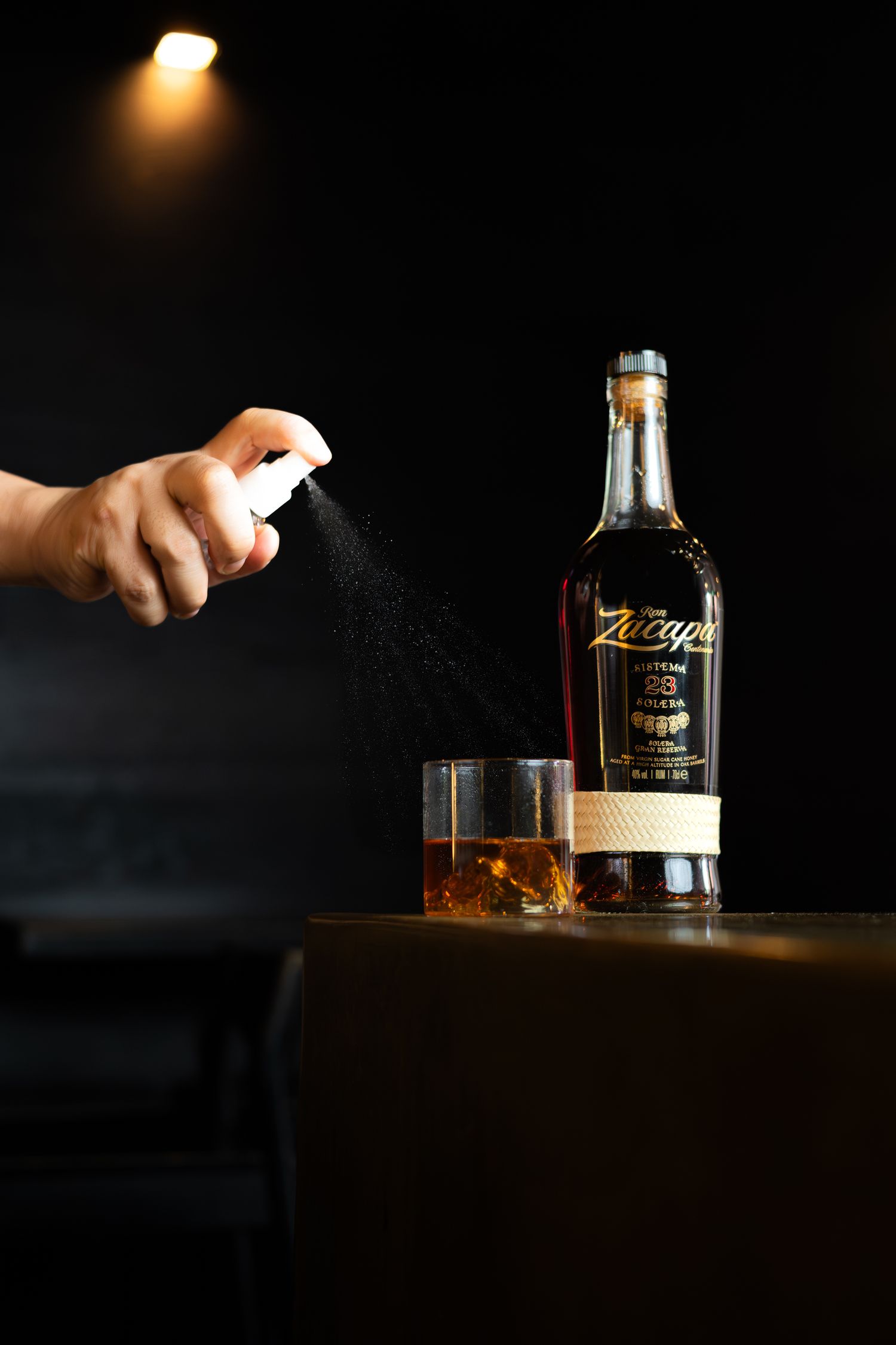 Hand spraying garnish over whiskey glass next to dark bottle in dramatic lighting.