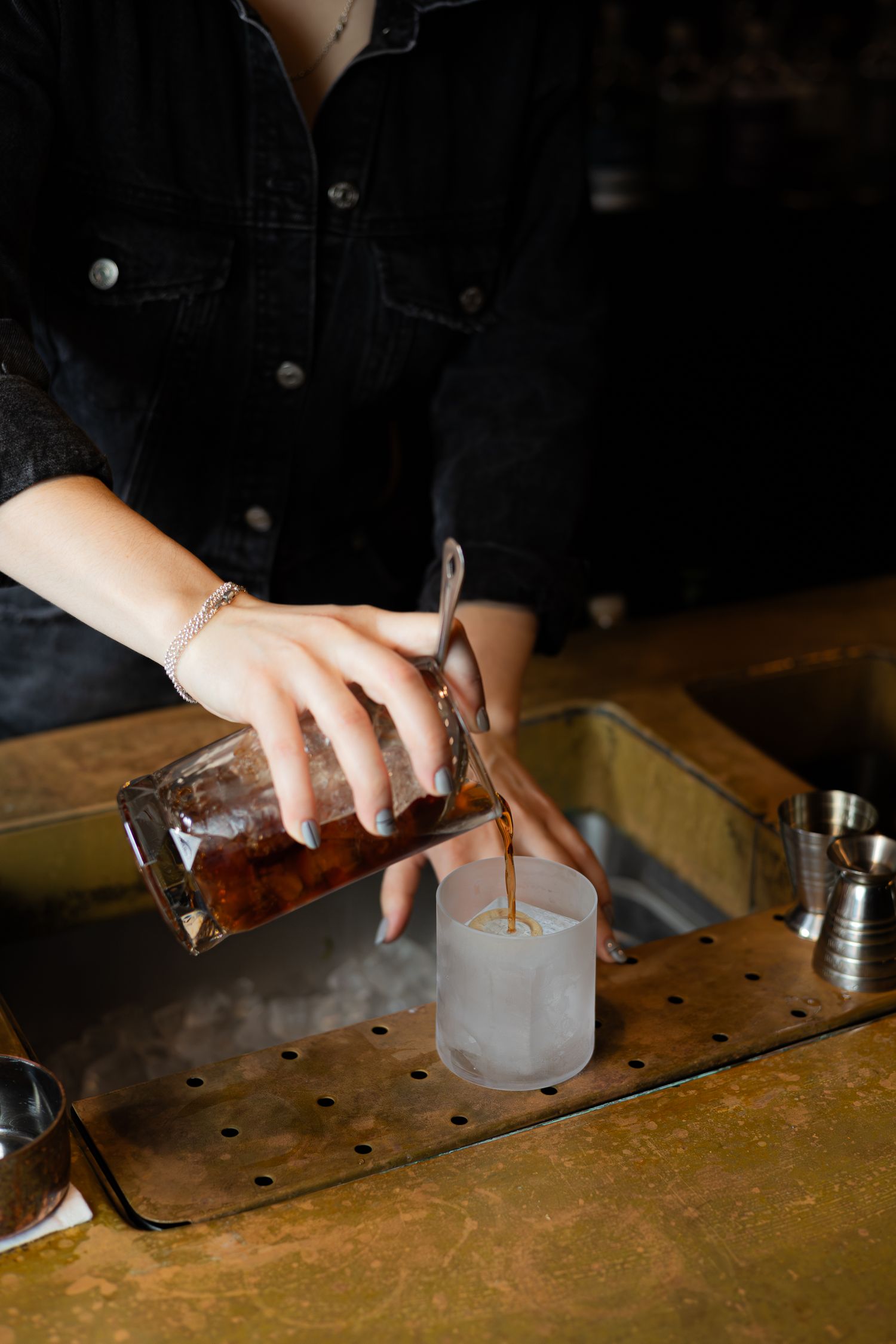 A bartender pours a dark liquid into a frosted glass on a wooden bar top.