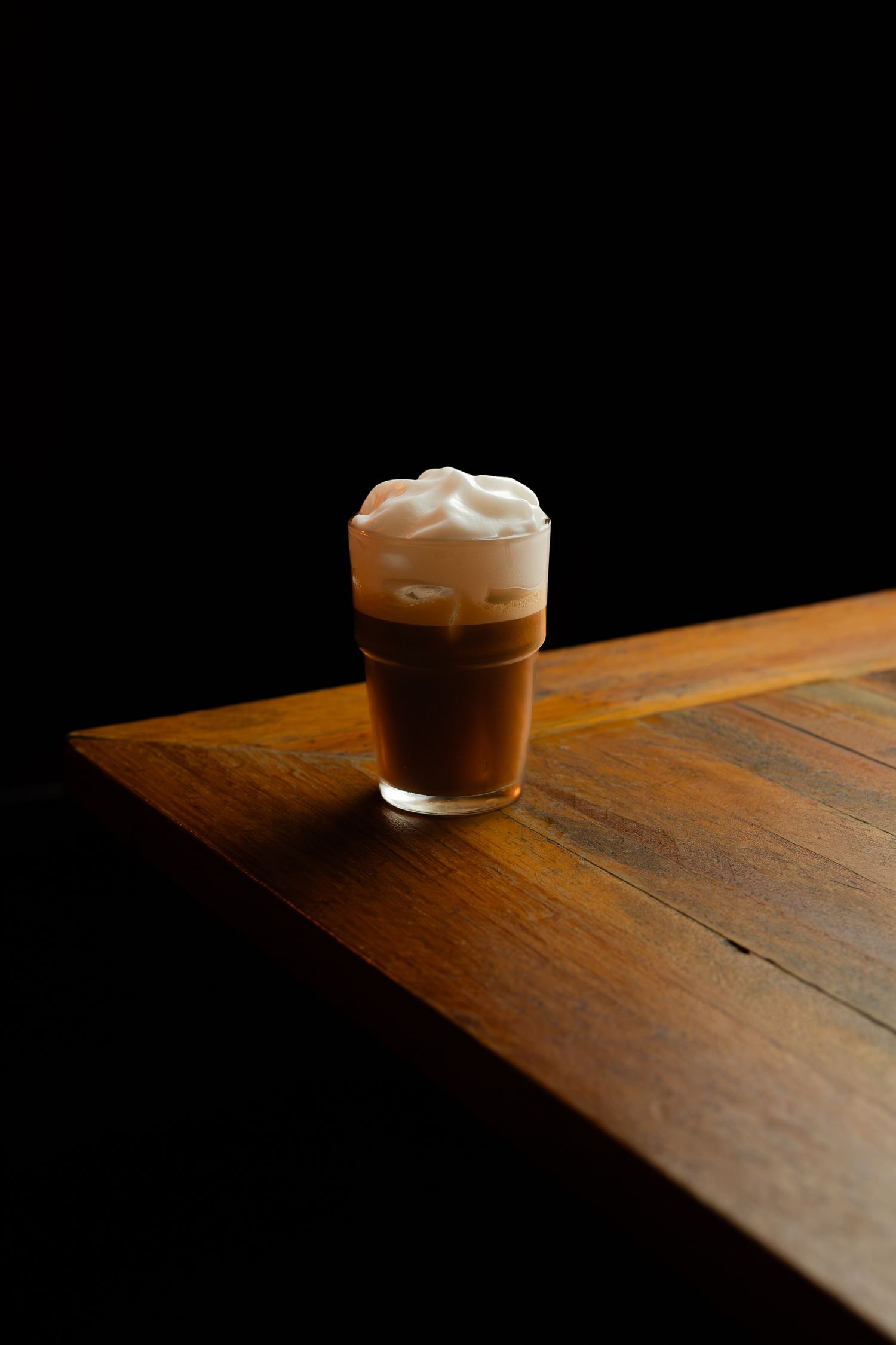 A layered coffee drink with whipped cream served in a glass on a wooden table corner.