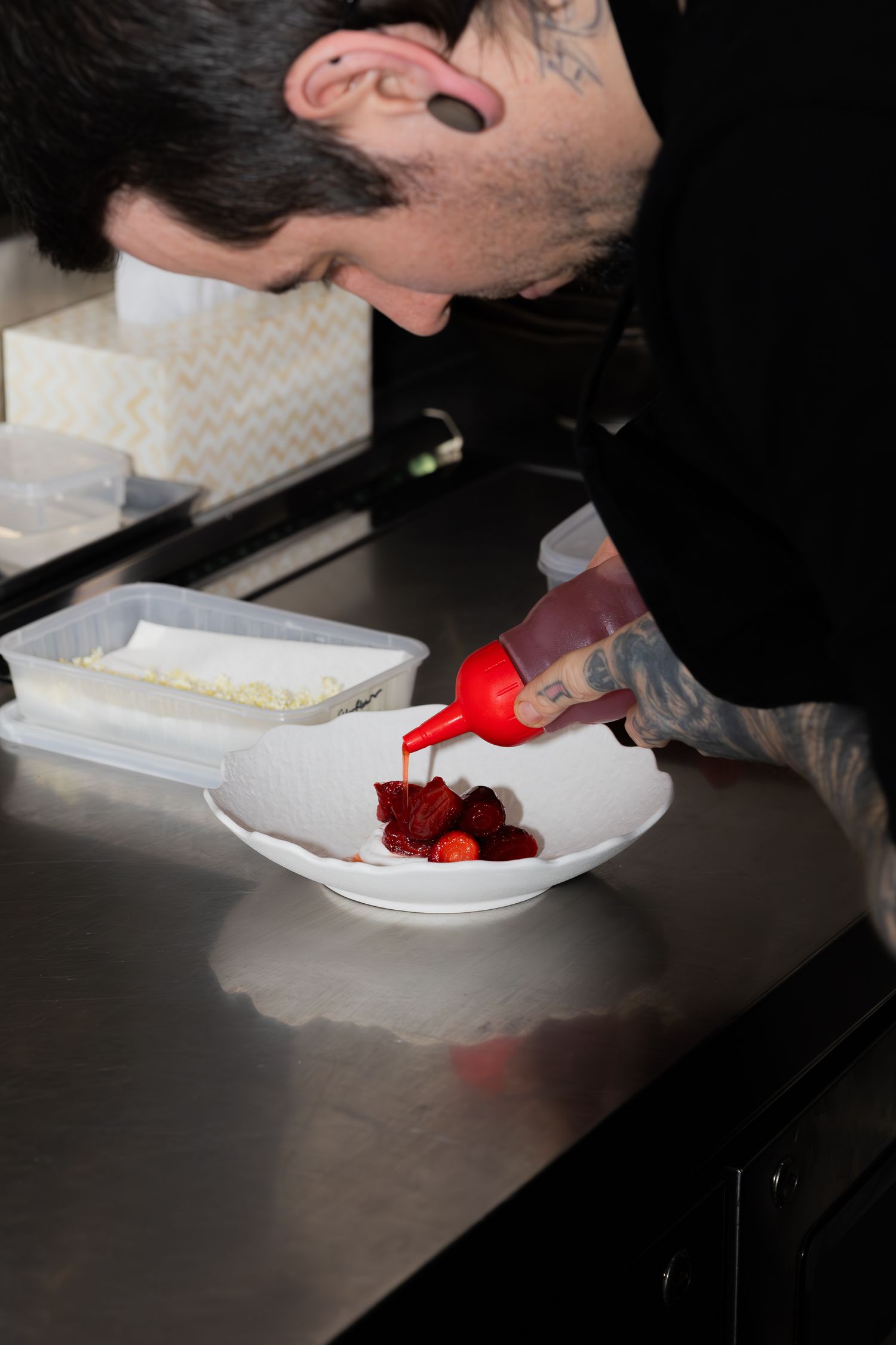 Chef preparing a plated dessert with red berries and various ingredients on a stainless steel counter.