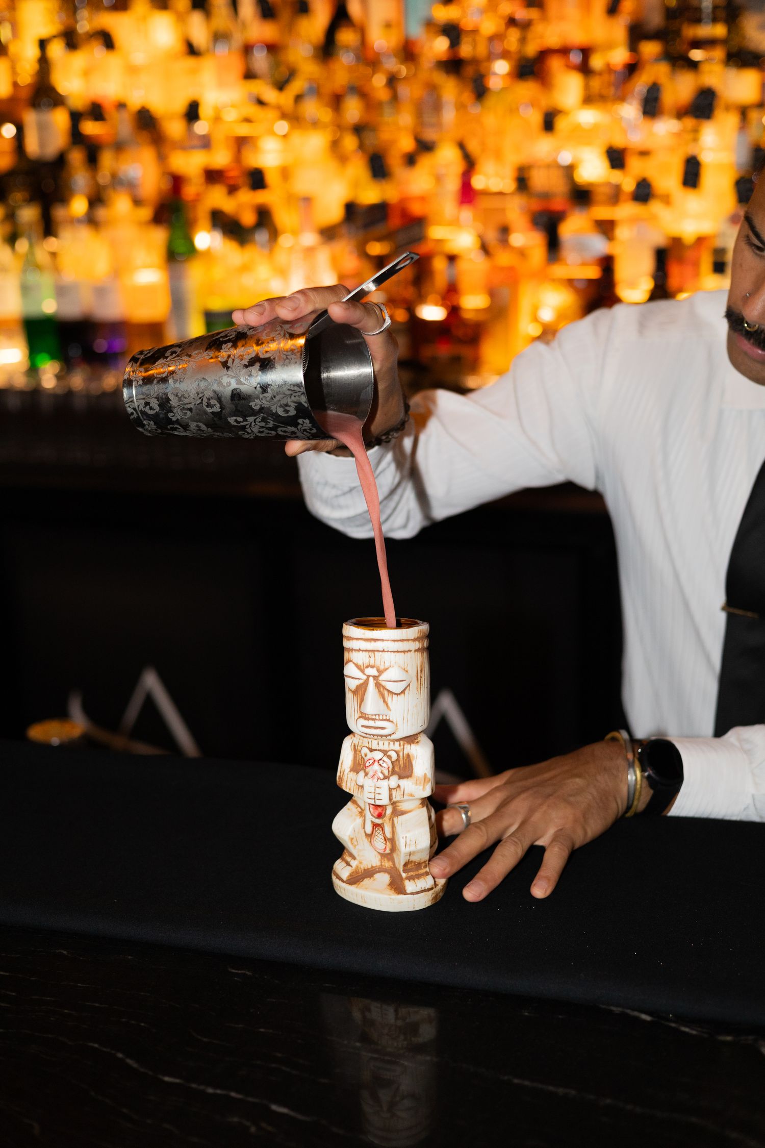 Cocktail being poured into decorative tiki mug at dimly lit bar.