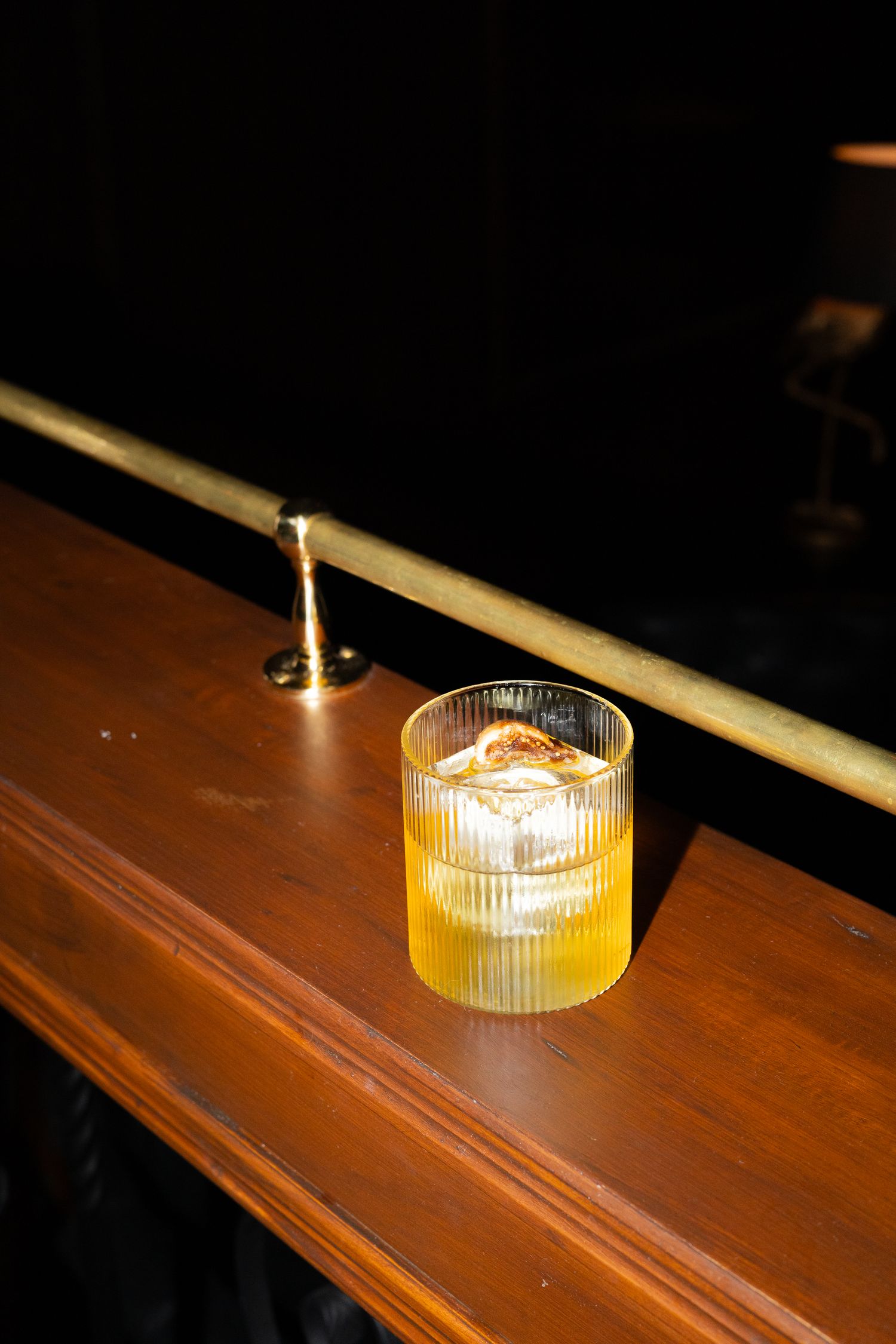 A glowing amber cocktail in a ribbed glass sits on a wooden bar rail with brass foot rail.