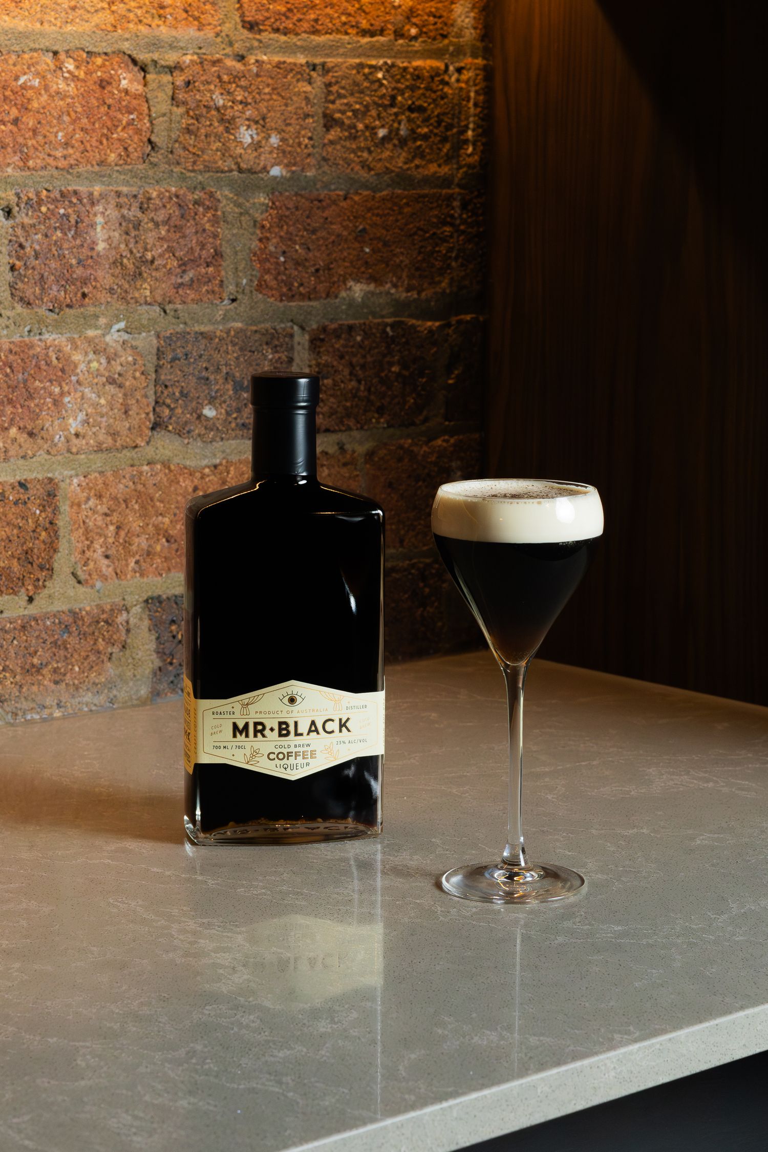 A black bottle of Mr Black liqueur next to an espresso martini on a marble counter.