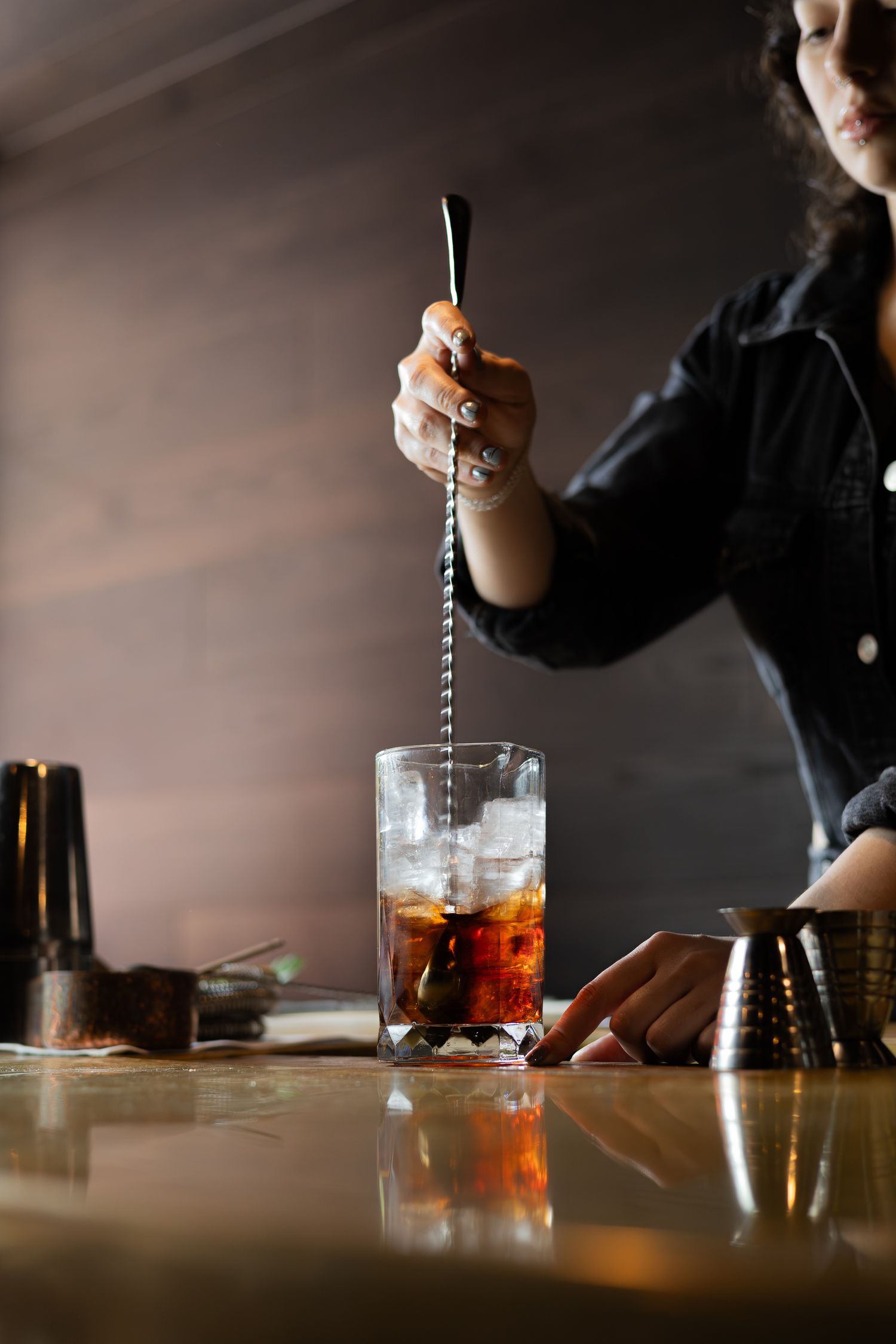 A mixologist stirs a cocktail with a long bar spoon in a tall glass filled with ice.