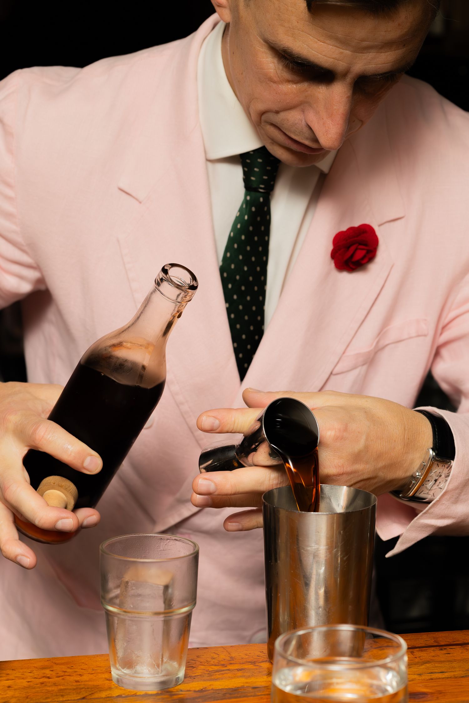 Bartender in pink blazer and red boutonniere mixing drinks at wooden bar counter.