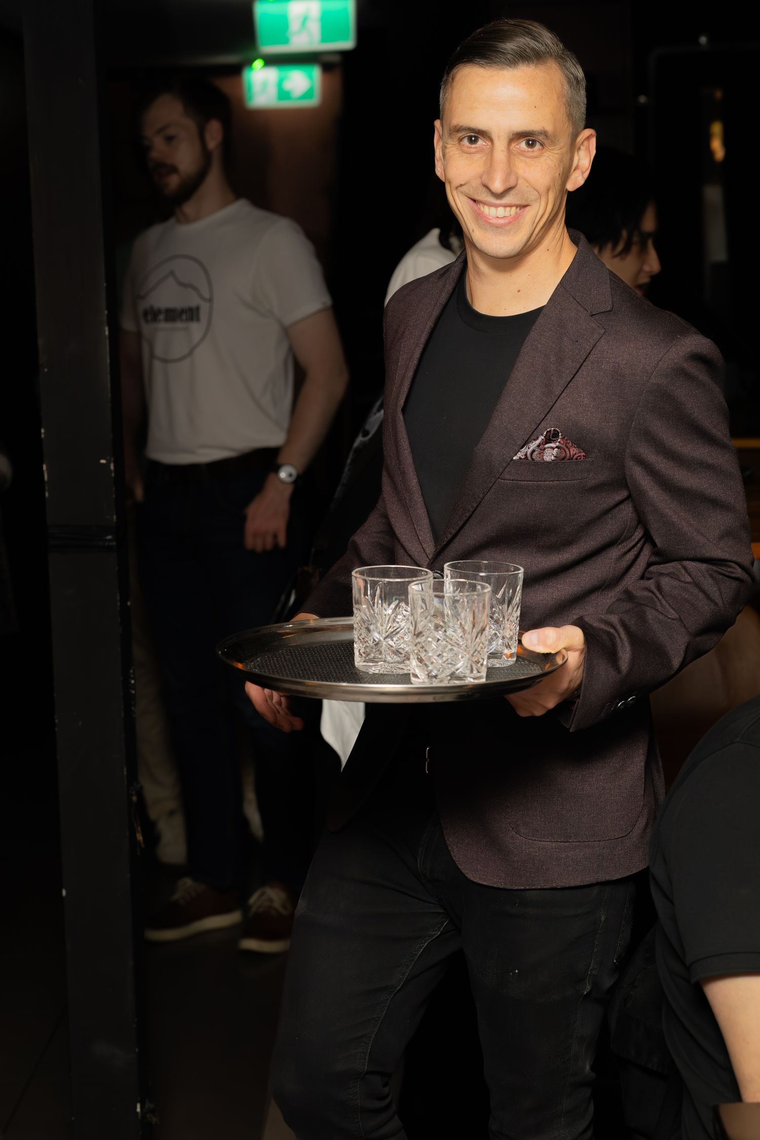 Server in blazer carrying crystal glasses on serving tray in dark venue.