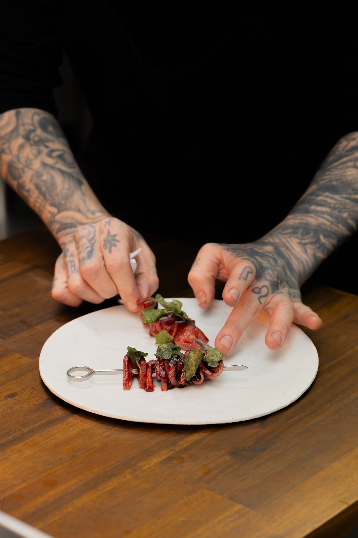 Tattooed hands plating a fine dining dish with red meat and microgreens on a white ceramic plate.