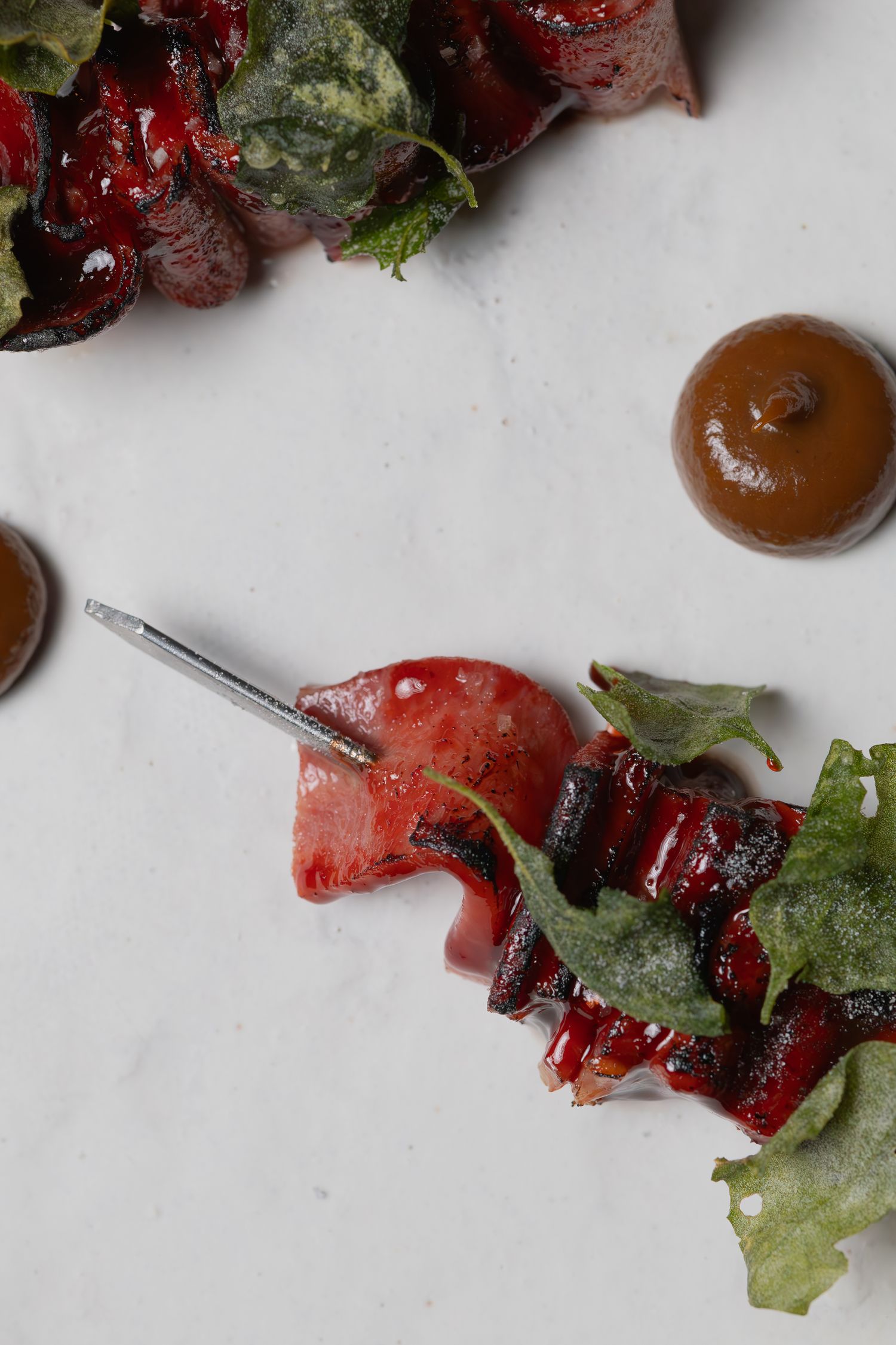Close-up of red meat skewer with green garnish and brown sauce dots on white surface.