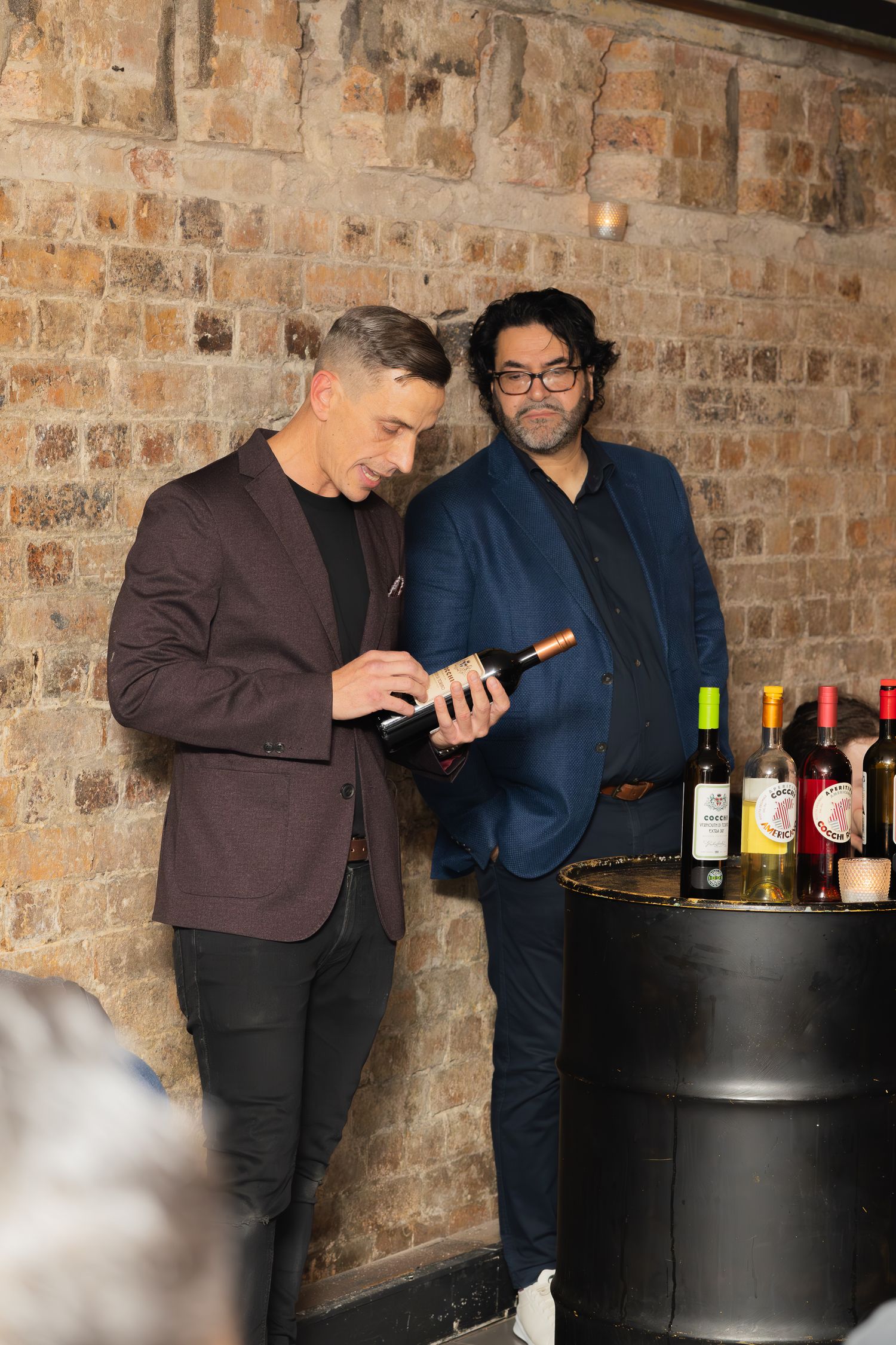 Two people in formal wear checking mobile device against brick wall near wine display.