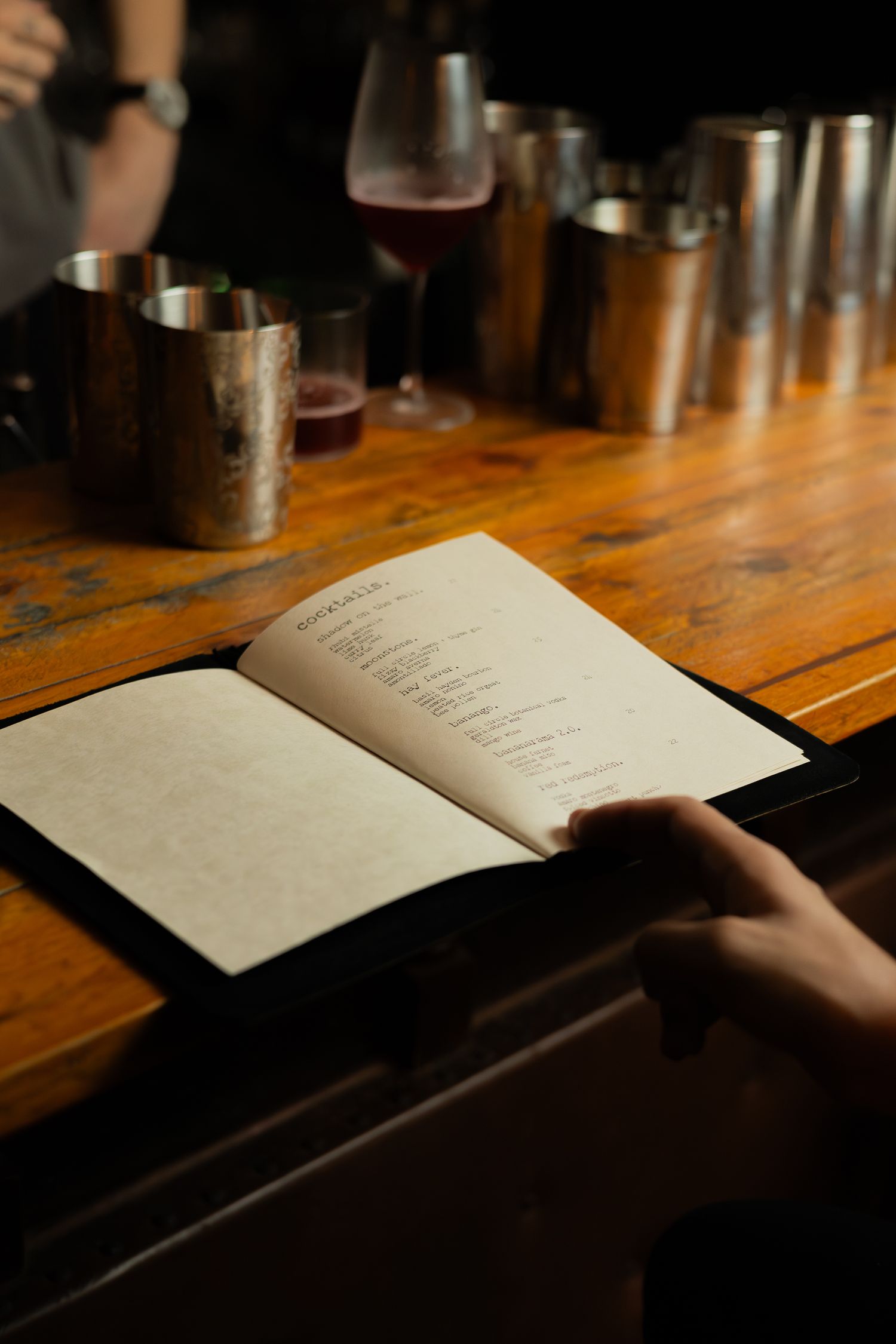 An open menu or book on a wooden bar counter with wine glasses in the background.