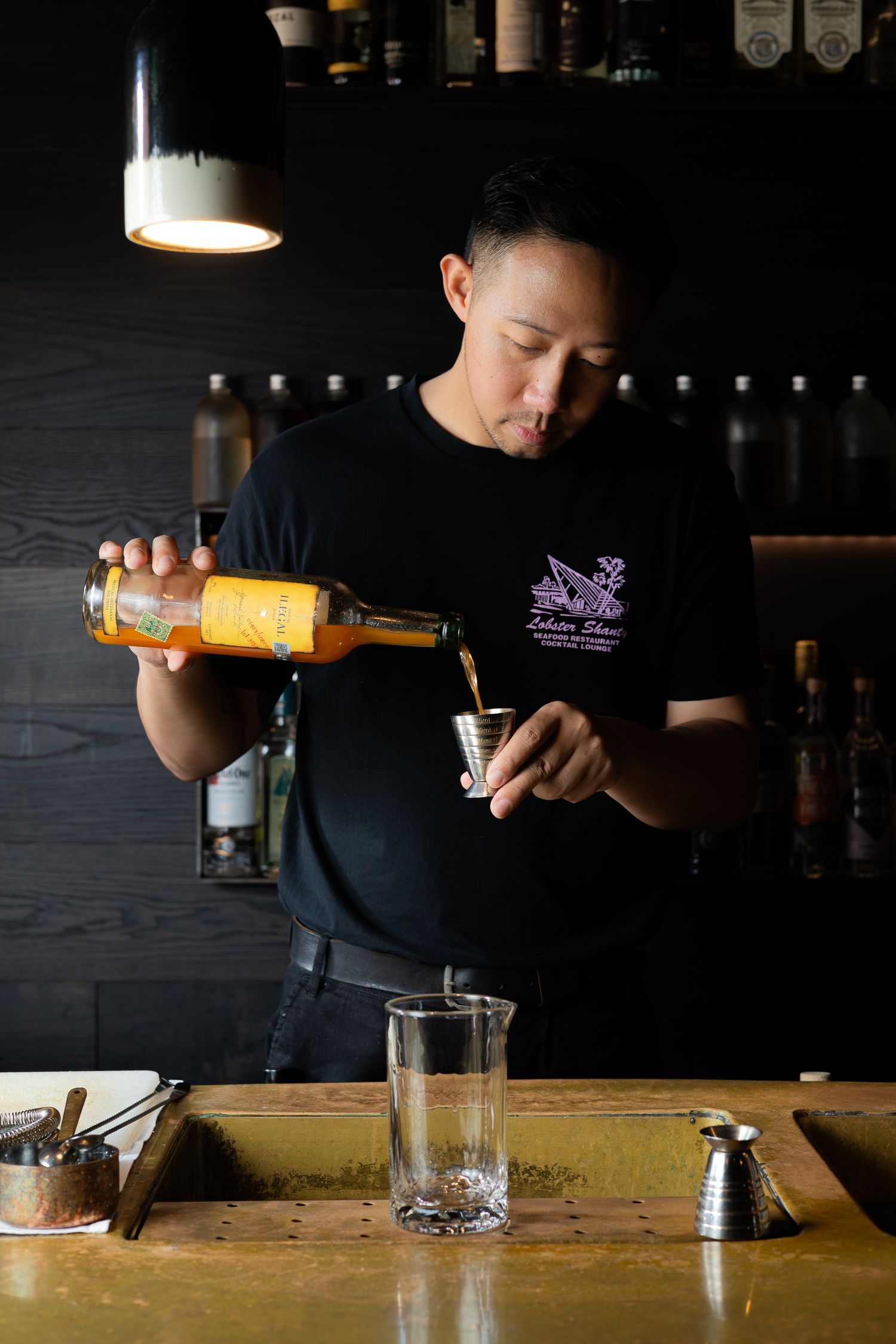 Bartender pouring orange liquid into mixing glass at bar.