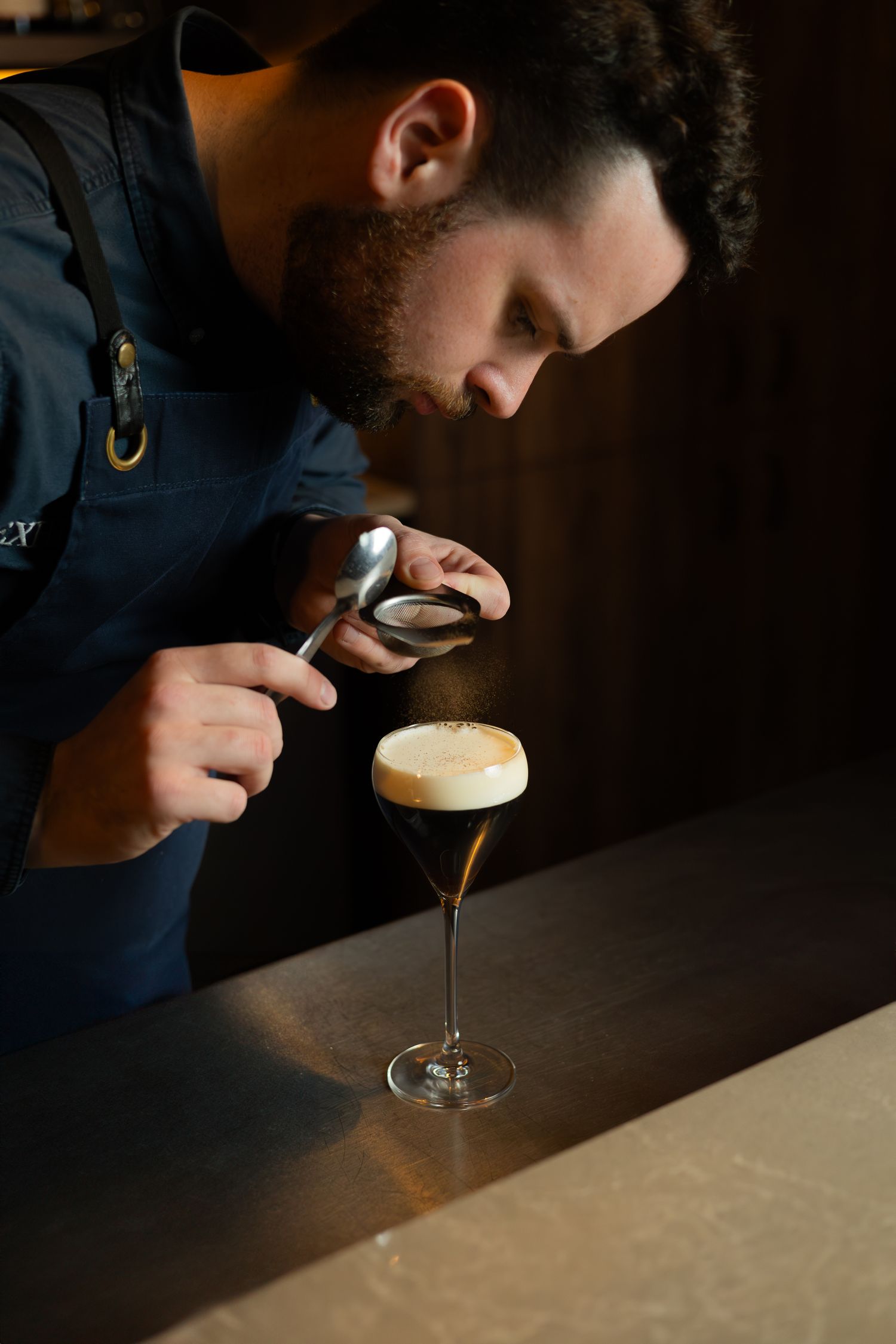 Barista examining espresso drink through magnifying glass in cocktail glass.