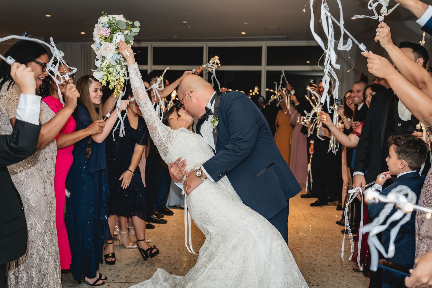 Bride and groom share romantic dip kiss at wedding reception exit with guests waving ribbon wands.