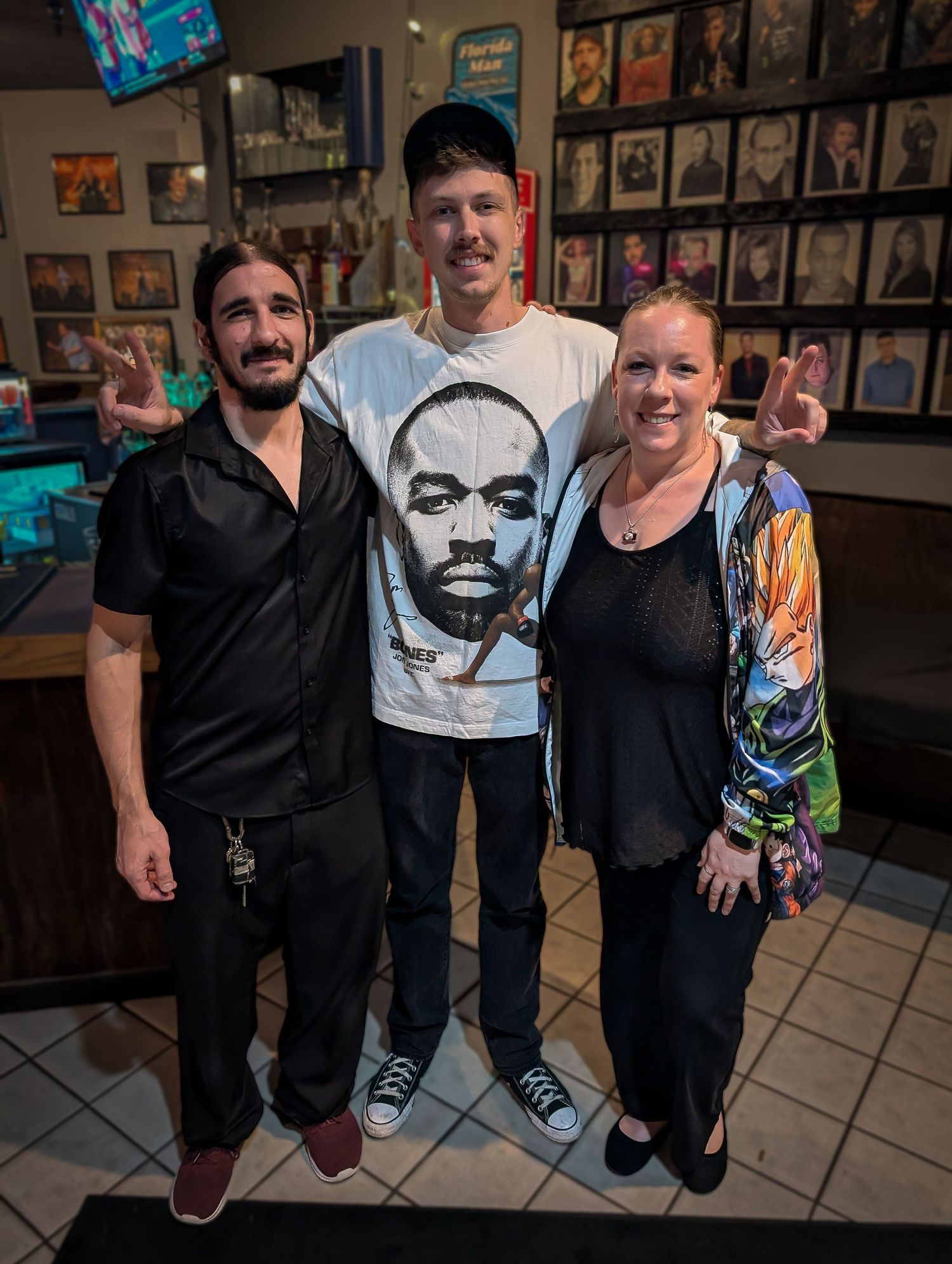 Cam Bertrand comedian. Three friends pose together smiling inside a restaurant, with the man in center wearing a graphic tee.