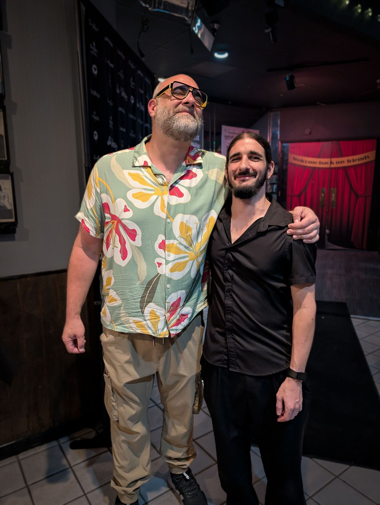 Kurt Metzger comedian. Two men pose together indoors, one tall in floral shirt and one shorter in black shirt at a venue.