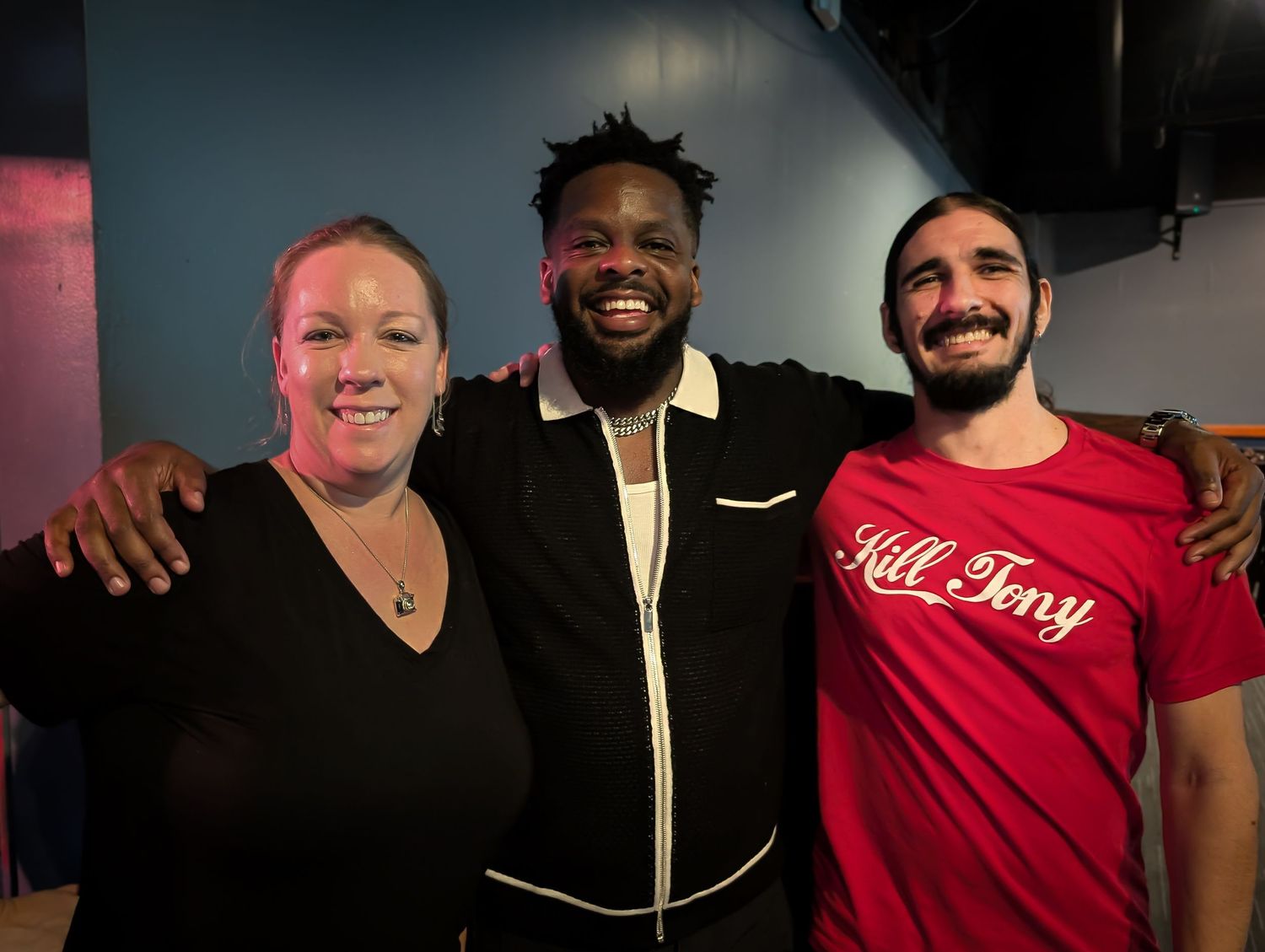 Dedrick Flynn. Three friends smiling together at a comedy club, woman on left, tall man in center, man in red Kill Tony shirt on right.